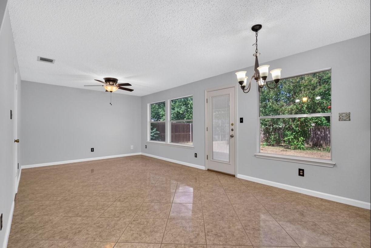 Empty tiled living area with a ceiling fan, chandelier, and windows overlooking a fenced yard.