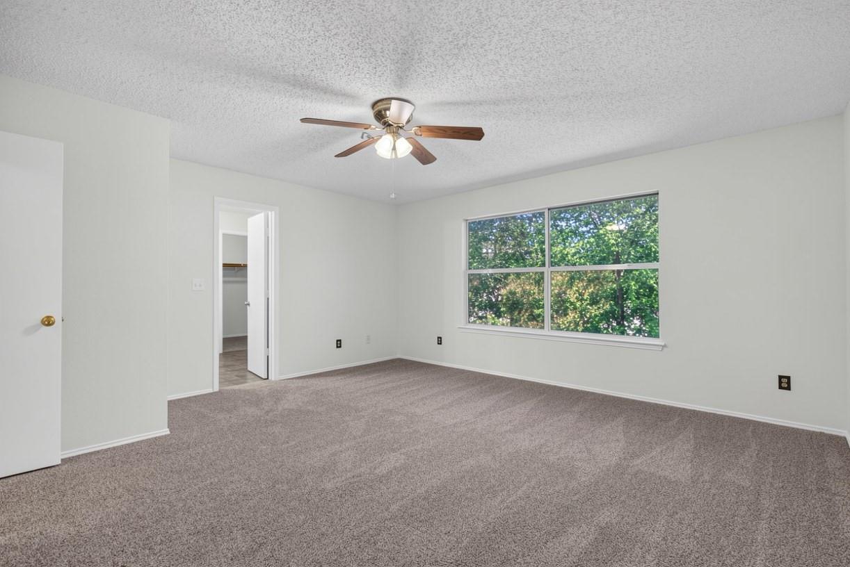 Empty carpeted bedroom with a ceiling fan and a large window overlooking leafy trees.