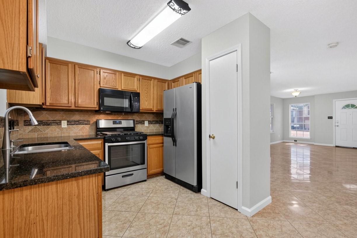 Kitchen with honey-colored cabinets, granite counters, stainless steel appliances, and an adjoining unfurnished living area.