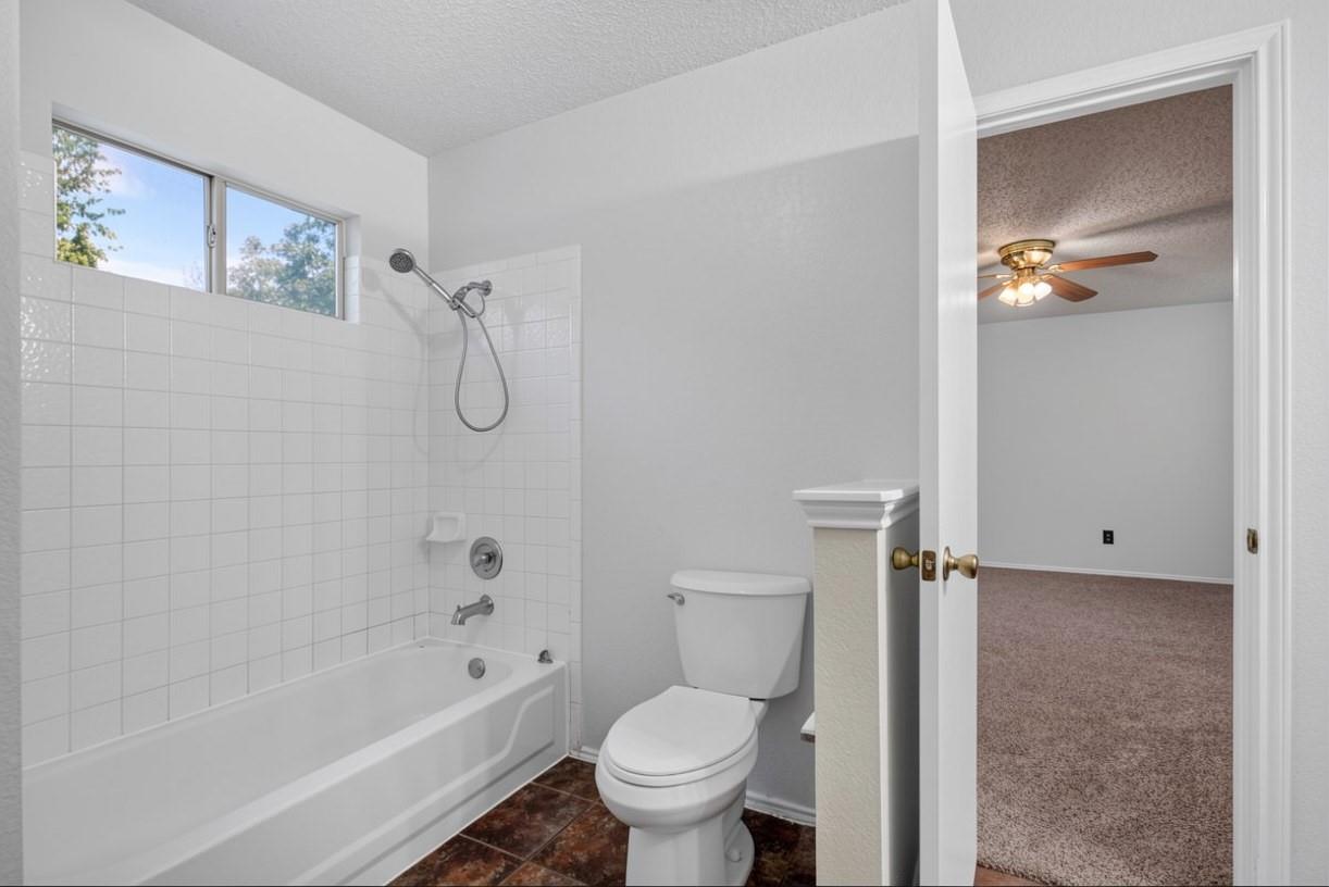 Bathroom with a white tub and shower beneath a high window, opening to an empty carpeted room.