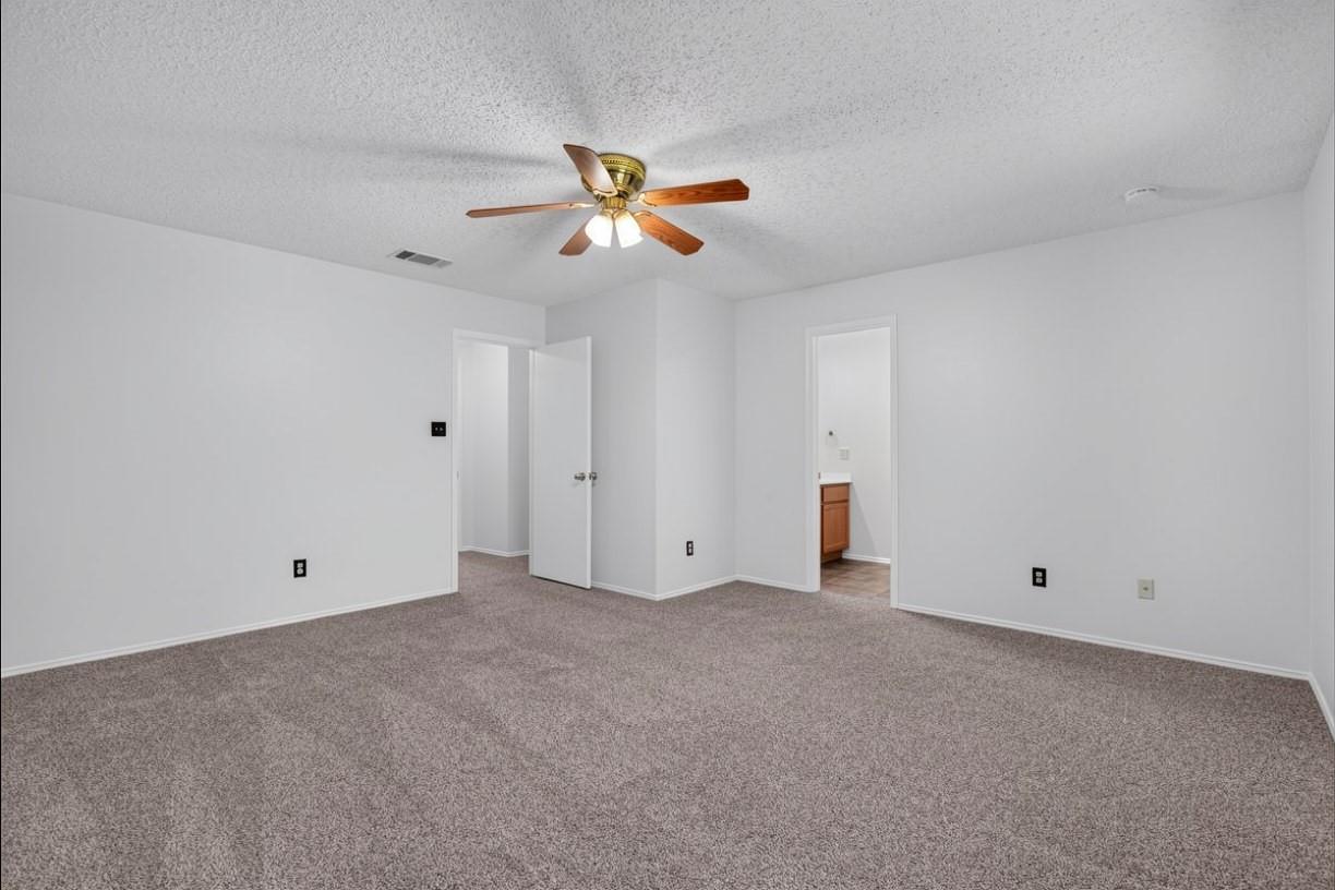 Empty bedroom with gray carpet and a wood-blade ceiling fan; an open doorway reveals part of a bathroom vanity.