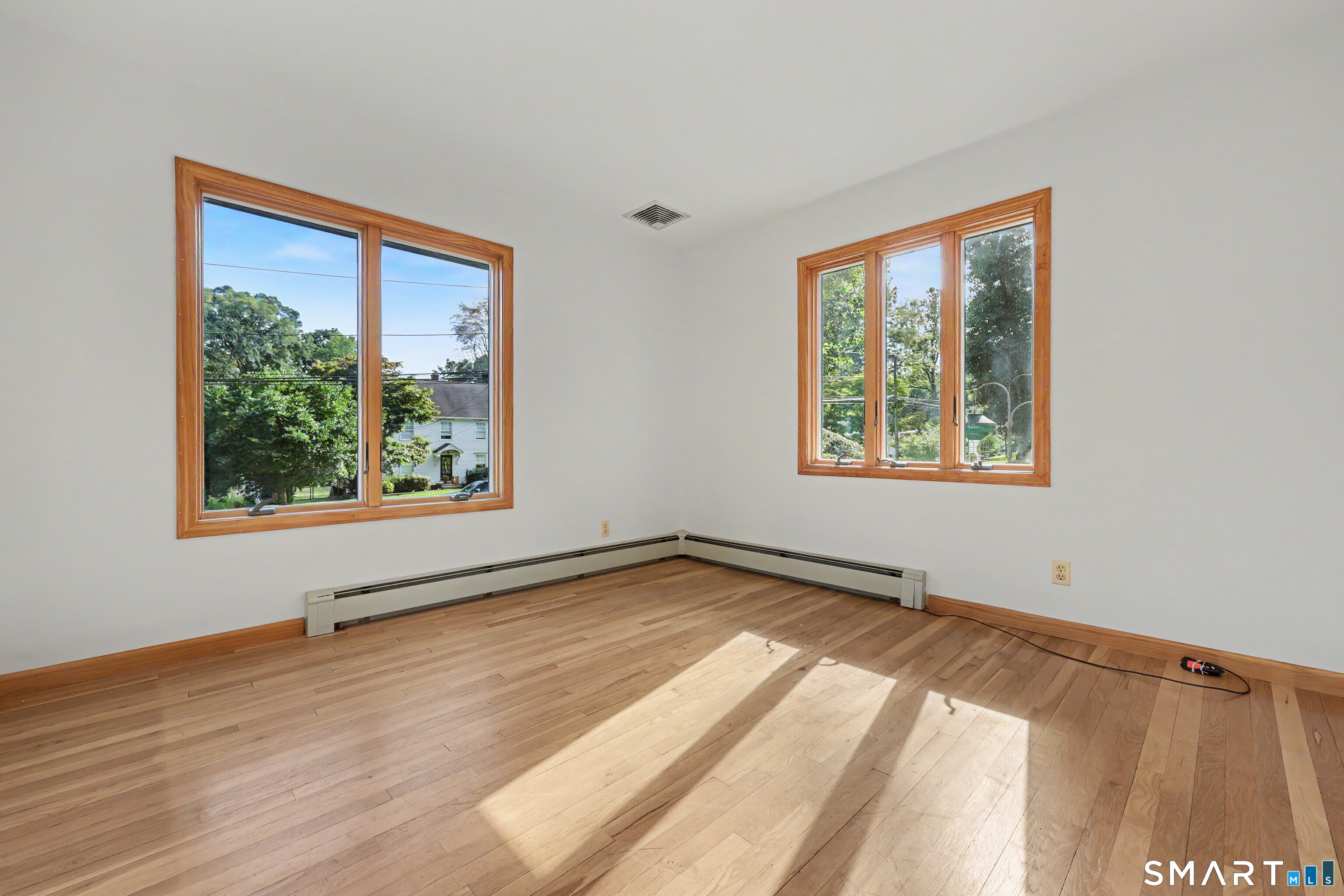 Empty room with hardwood floors, two wood-trimmed windows, and sunlight across the floor.