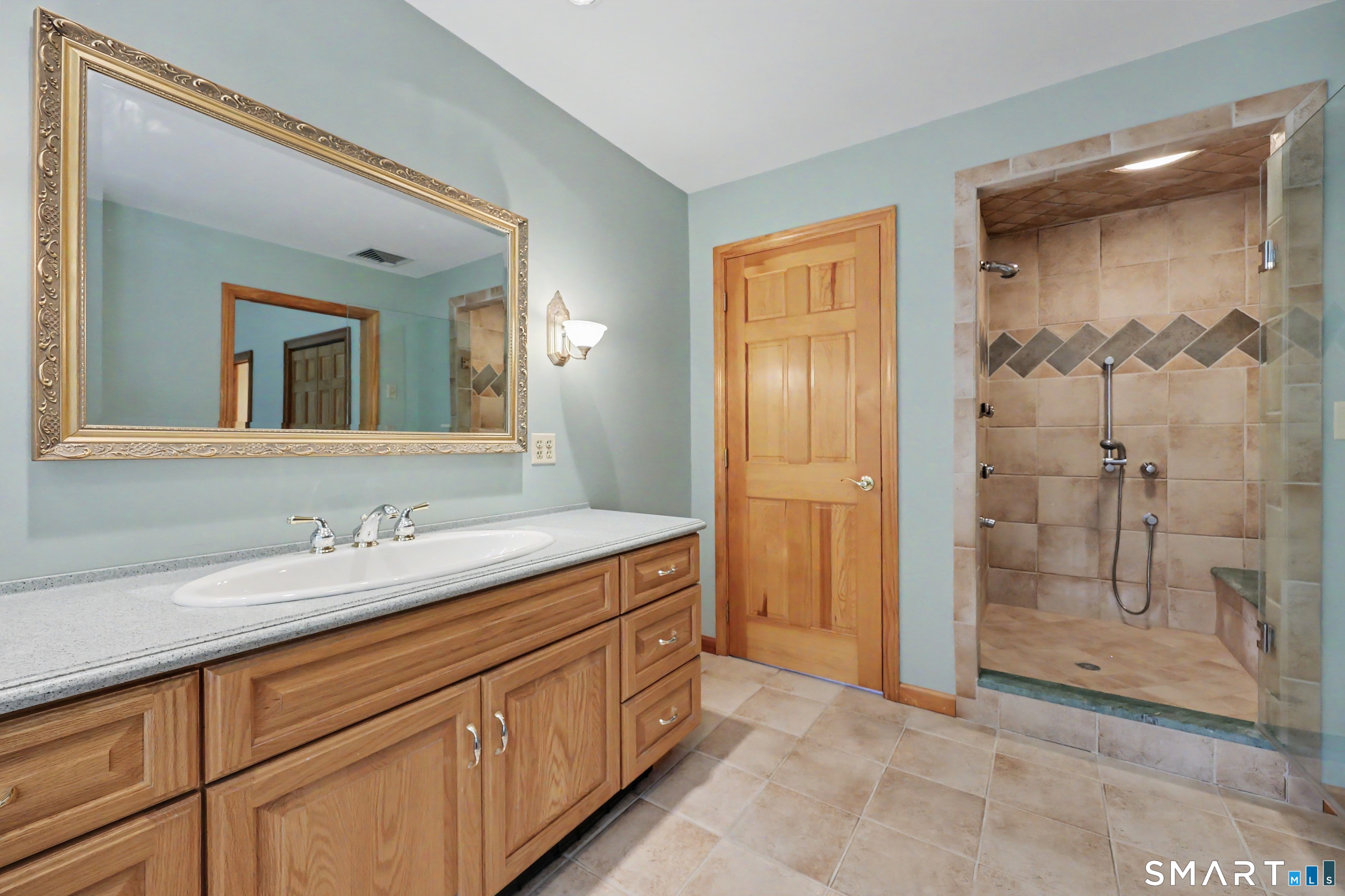 Bathroom with a wood vanity and ornate mirror beside a spacious tan-tiled walk-in shower.