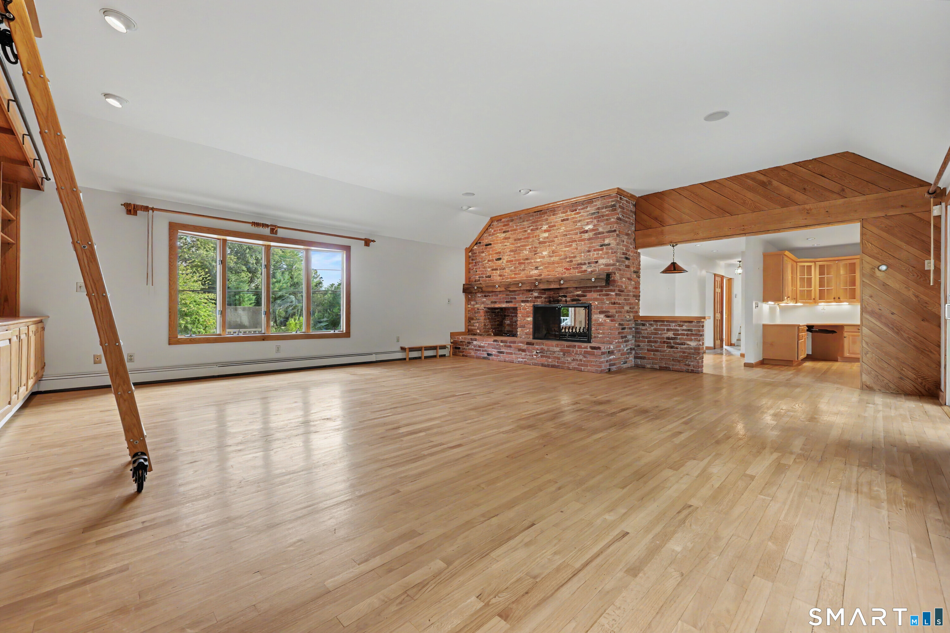 Vacant living room with hardwood floors, a brick fireplace, exposed wood framing, and an open kitchen.