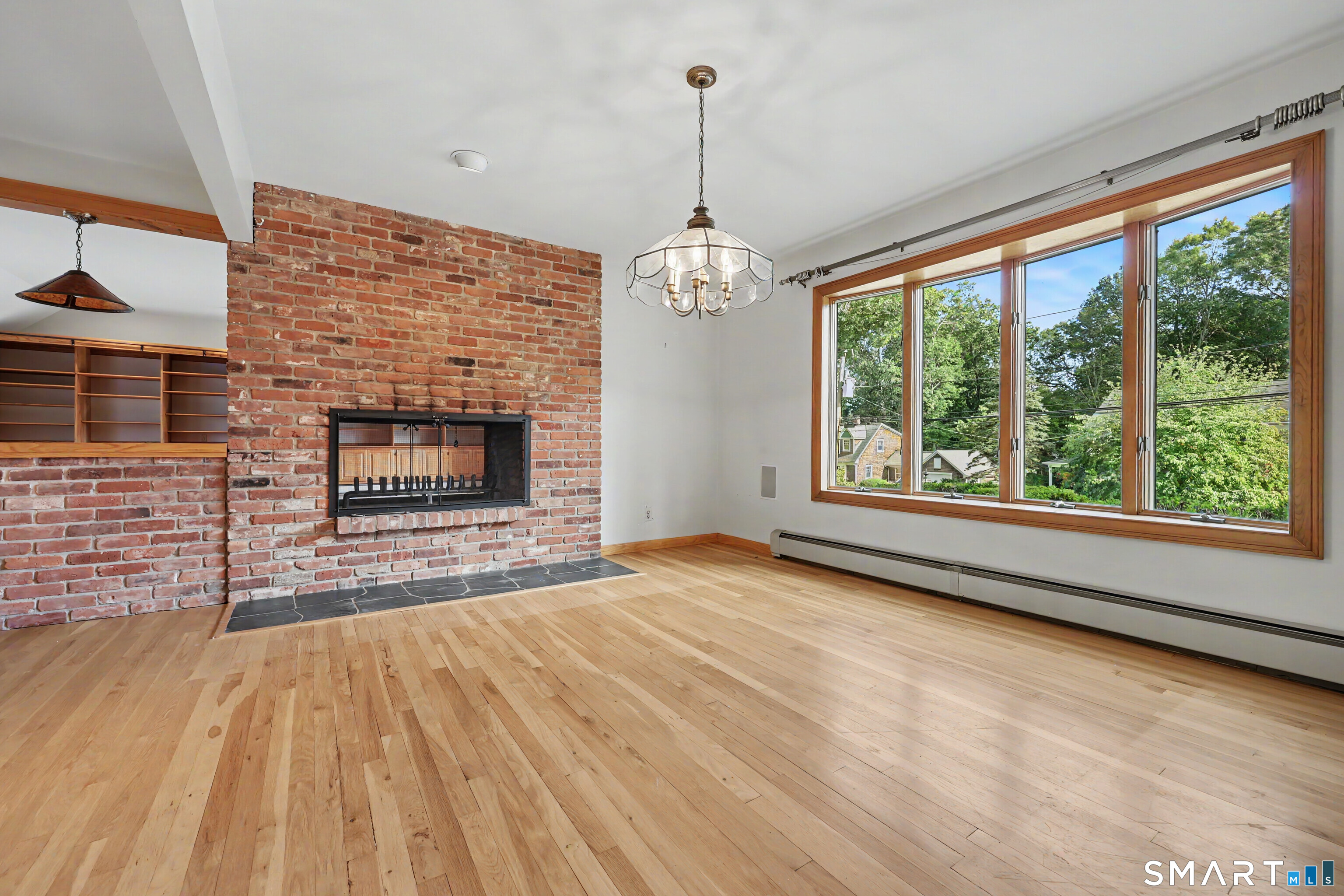 Empty living room with a brick fireplace, hardwood floors, and a wide window overlooking trees.