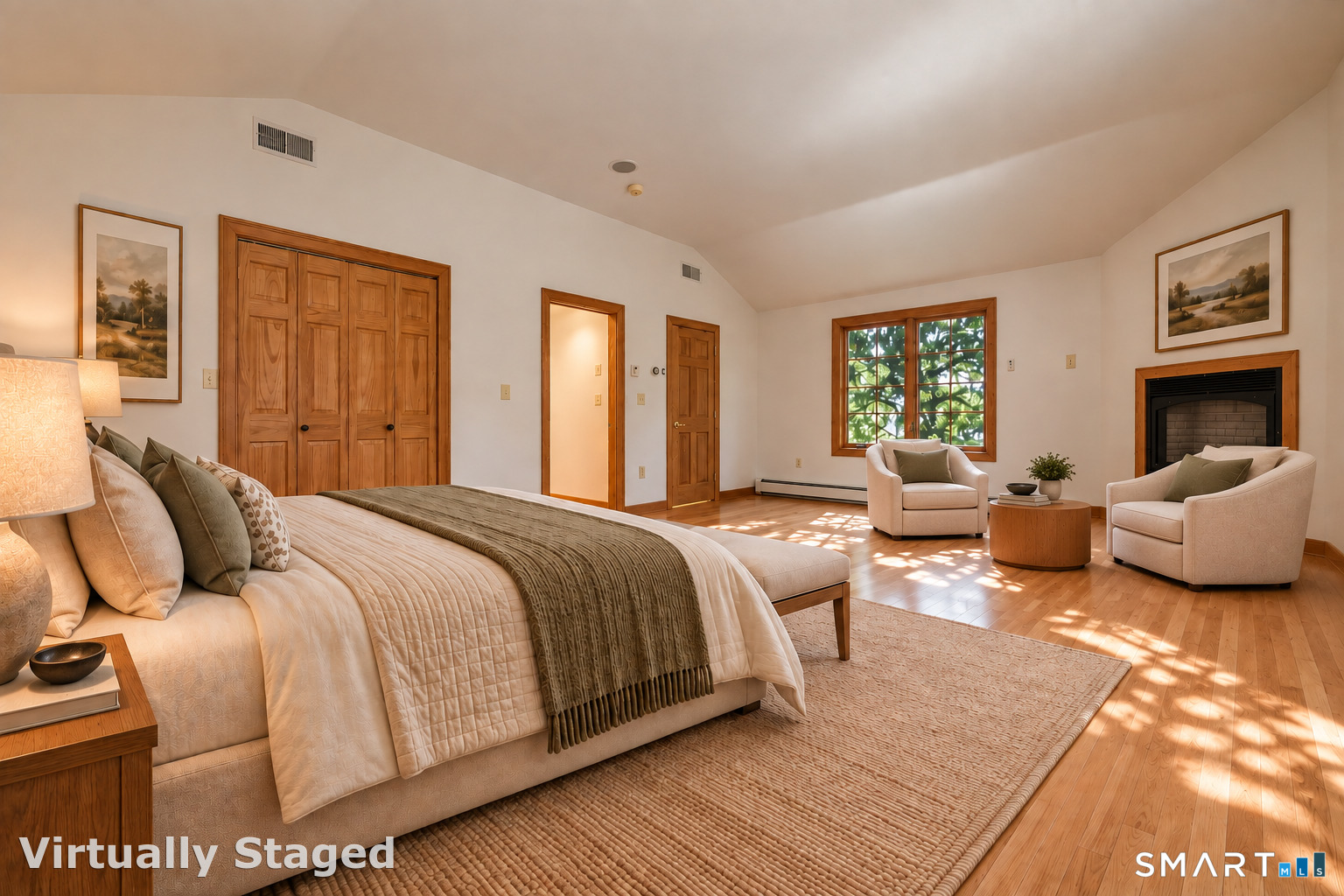 Bedroom with a large bed, two armchairs by a fireplace, and sunlight across the wood floor.