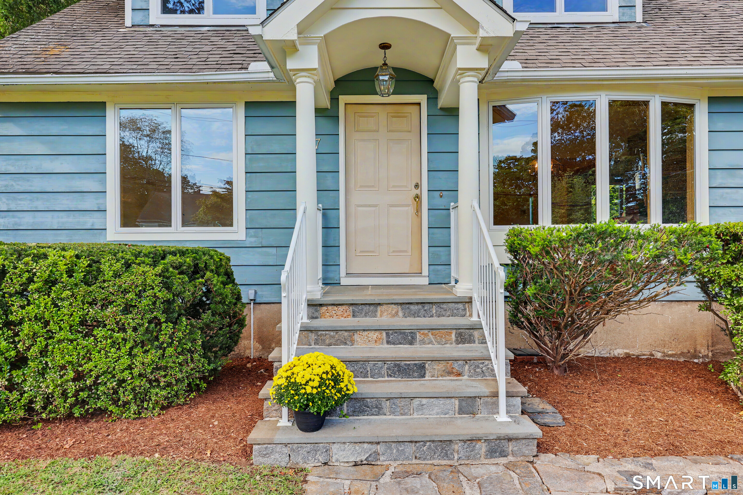 Blue house entrance with white porch columns, stone steps, and a potted yellow flowering plant.