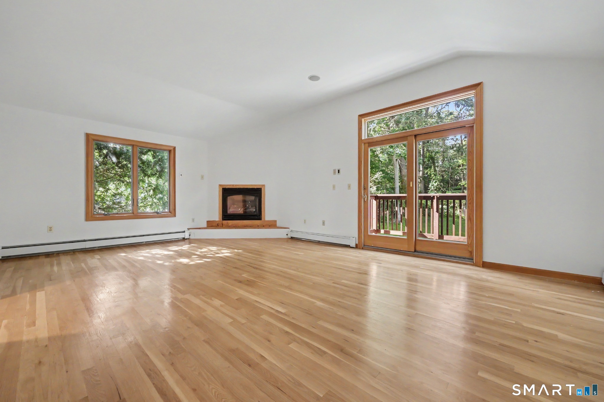 Vacant living room with hardwood floors, corner fireplace, and glass doors to a wooded deck.