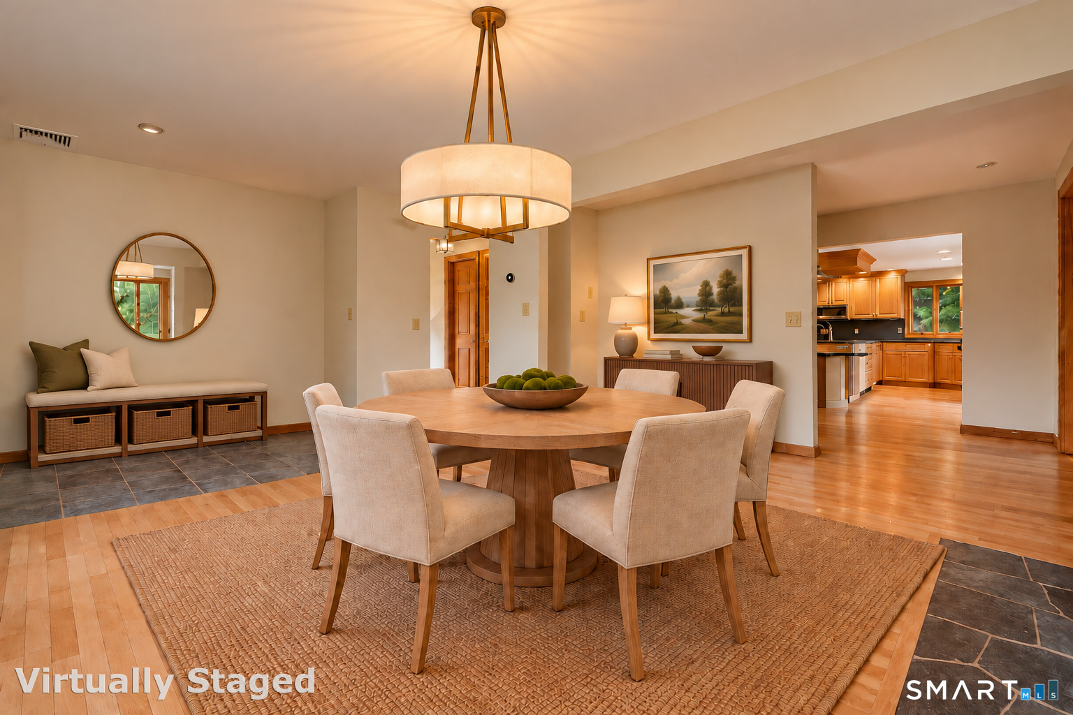 Dining room with a round wooden table, upholstered chairs, pendant light, and adjoining kitchen.