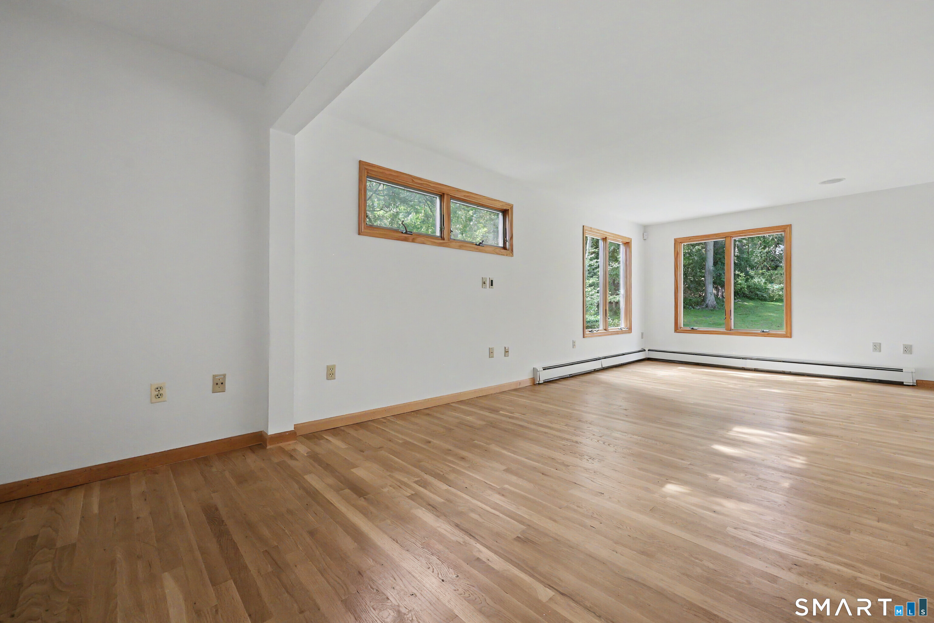 Empty room with wide-plank hardwood floors and wood-trimmed windows overlooking trees.