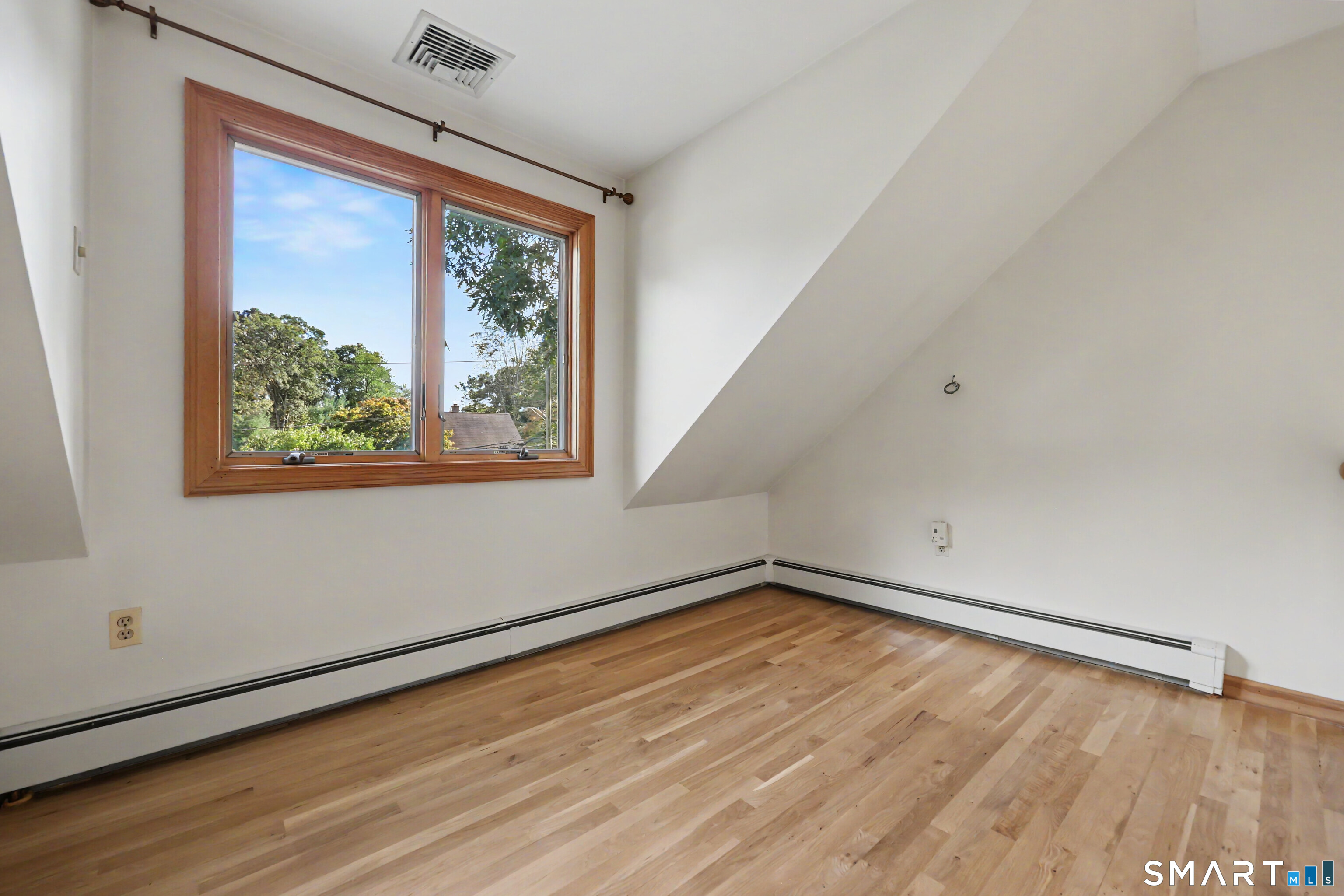 Vacant upstairs bedroom with hardwood floors, a sloped ceiling, and a large wood-trimmed window.