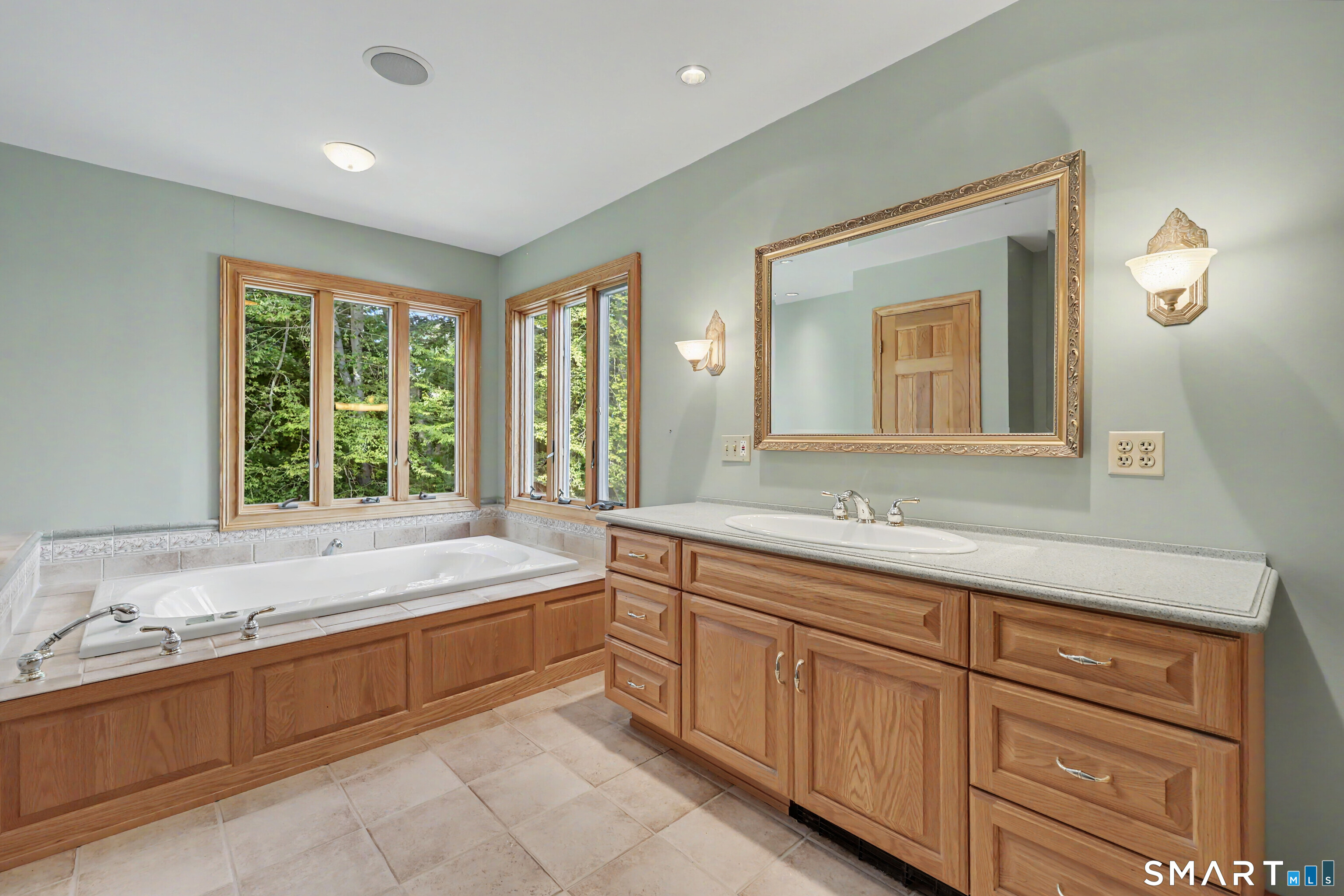 Bathroom with corner soaking tub, wood vanity, and windows overlooking greenery.