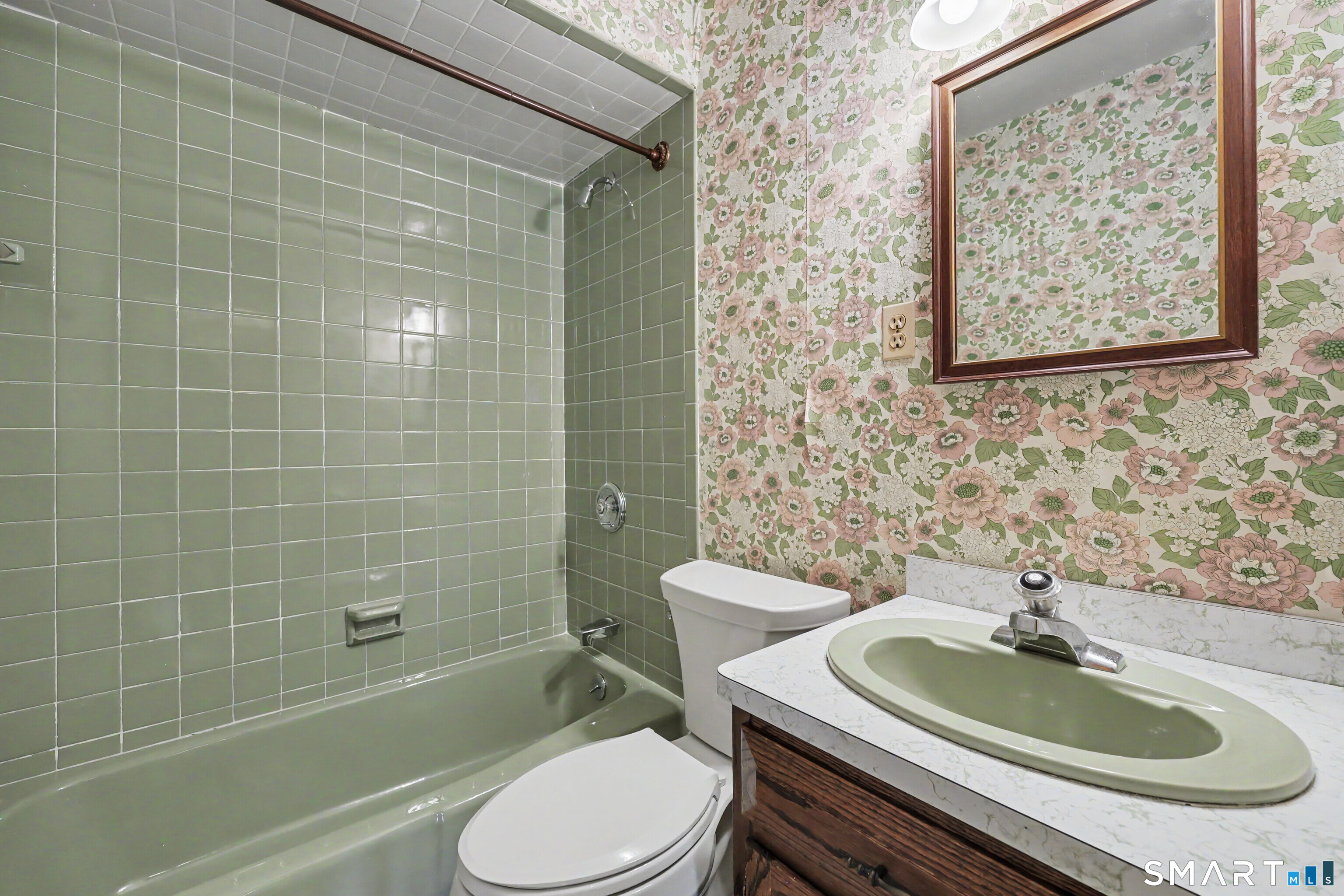 Bathroom with pale green tiled tub and shower, floral wallpaper, and green sink on a wood vanity.
