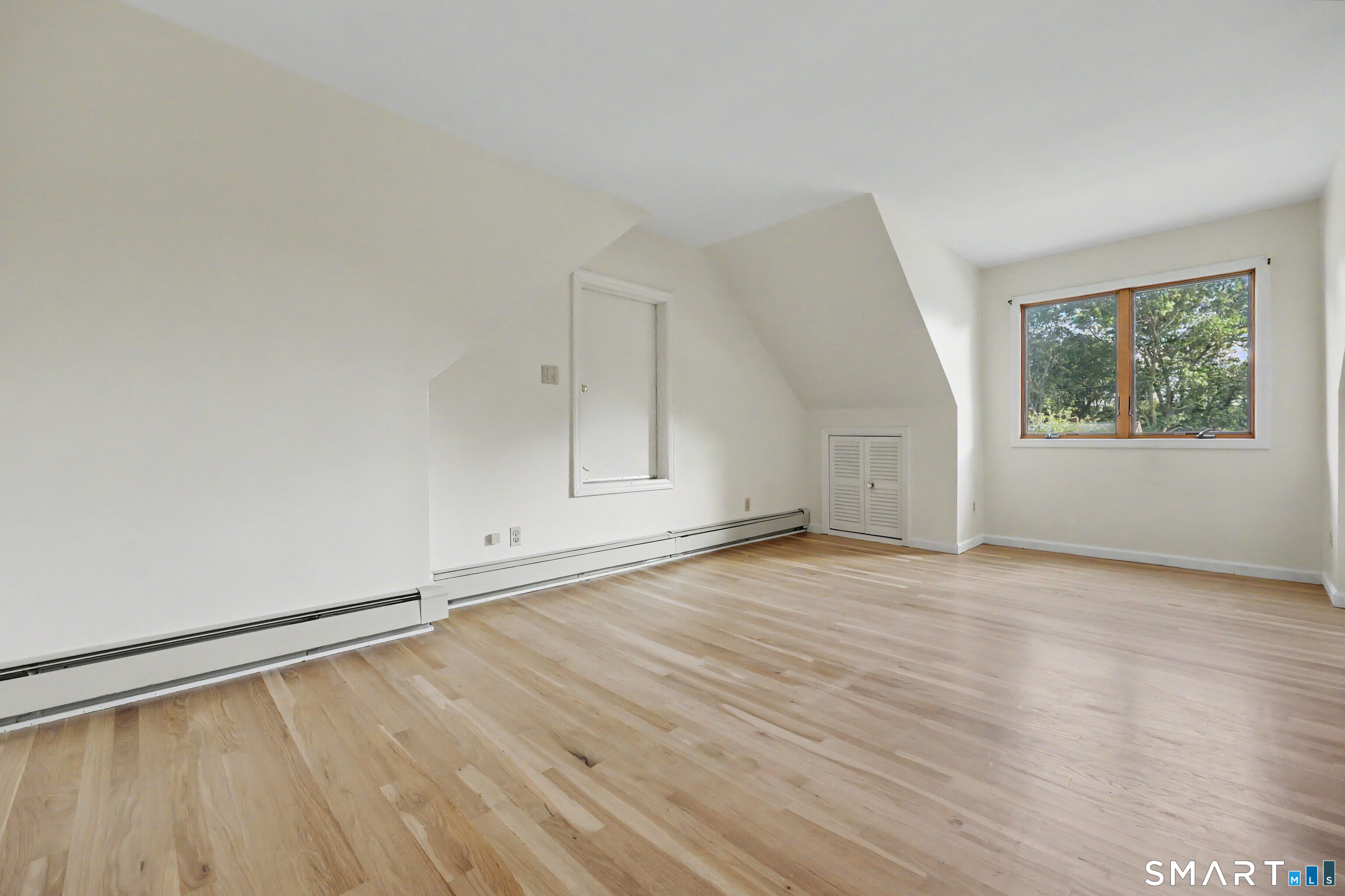 Vacant room with light hardwood floors, a sloped ceiling, and a window overlooking trees.