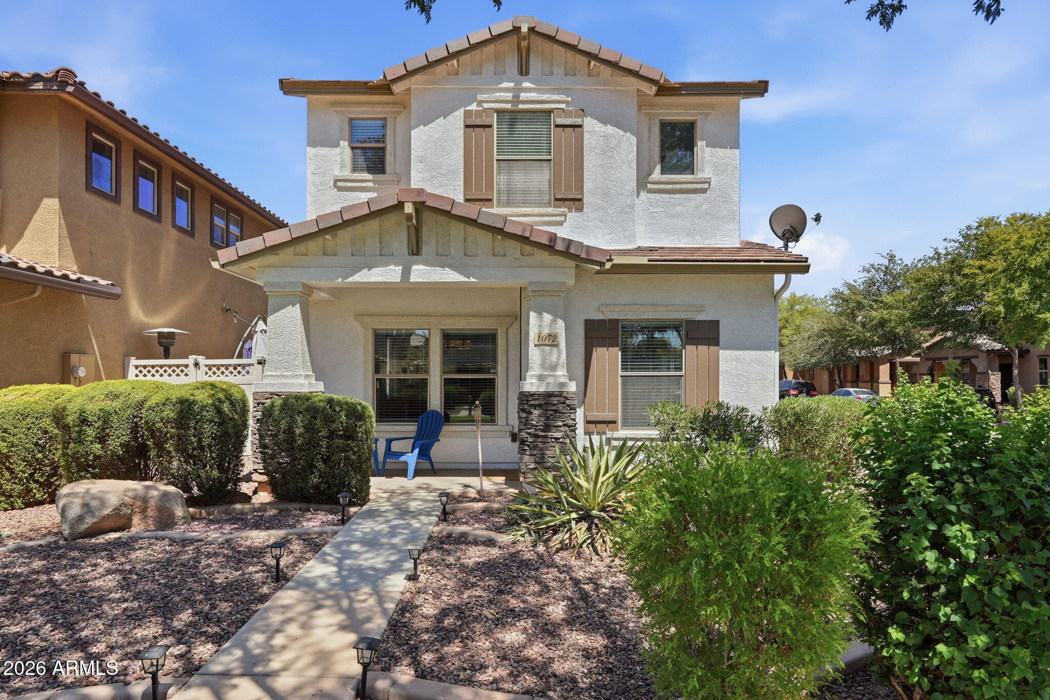 Two-story stucco house with brown shutters, a blue porch chair, and desert landscaping.