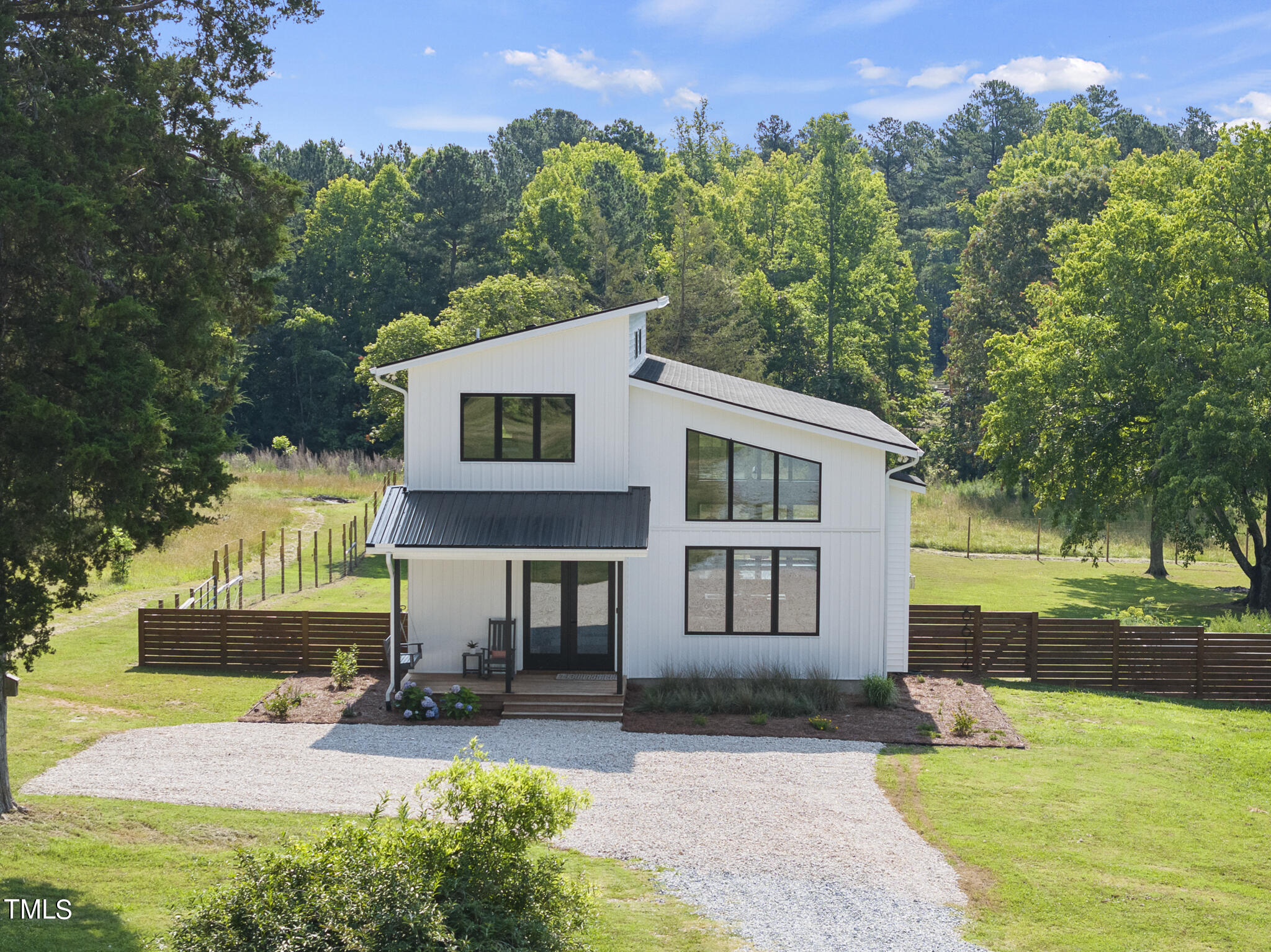 Modern white two-story home with large windows, a gravel driveway, and a fenced, wooded yard.