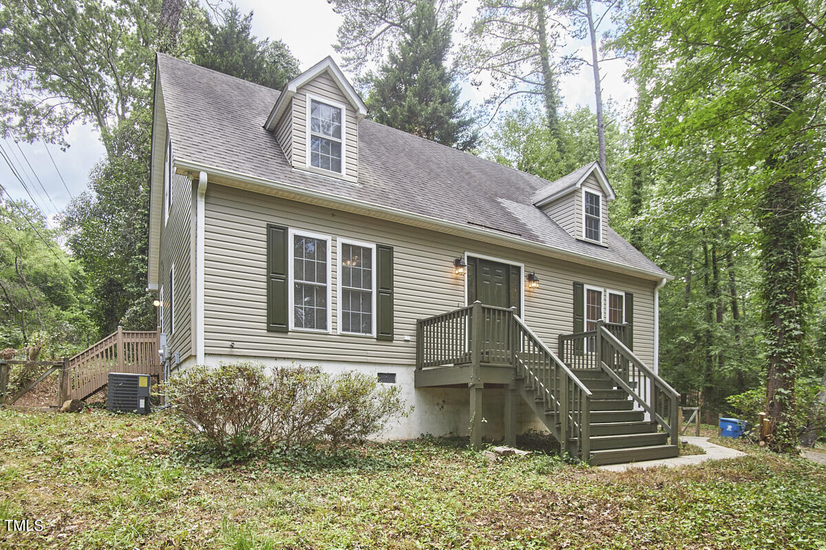 Beige-sided house with two dormers and a raised entry deck among mature trees.