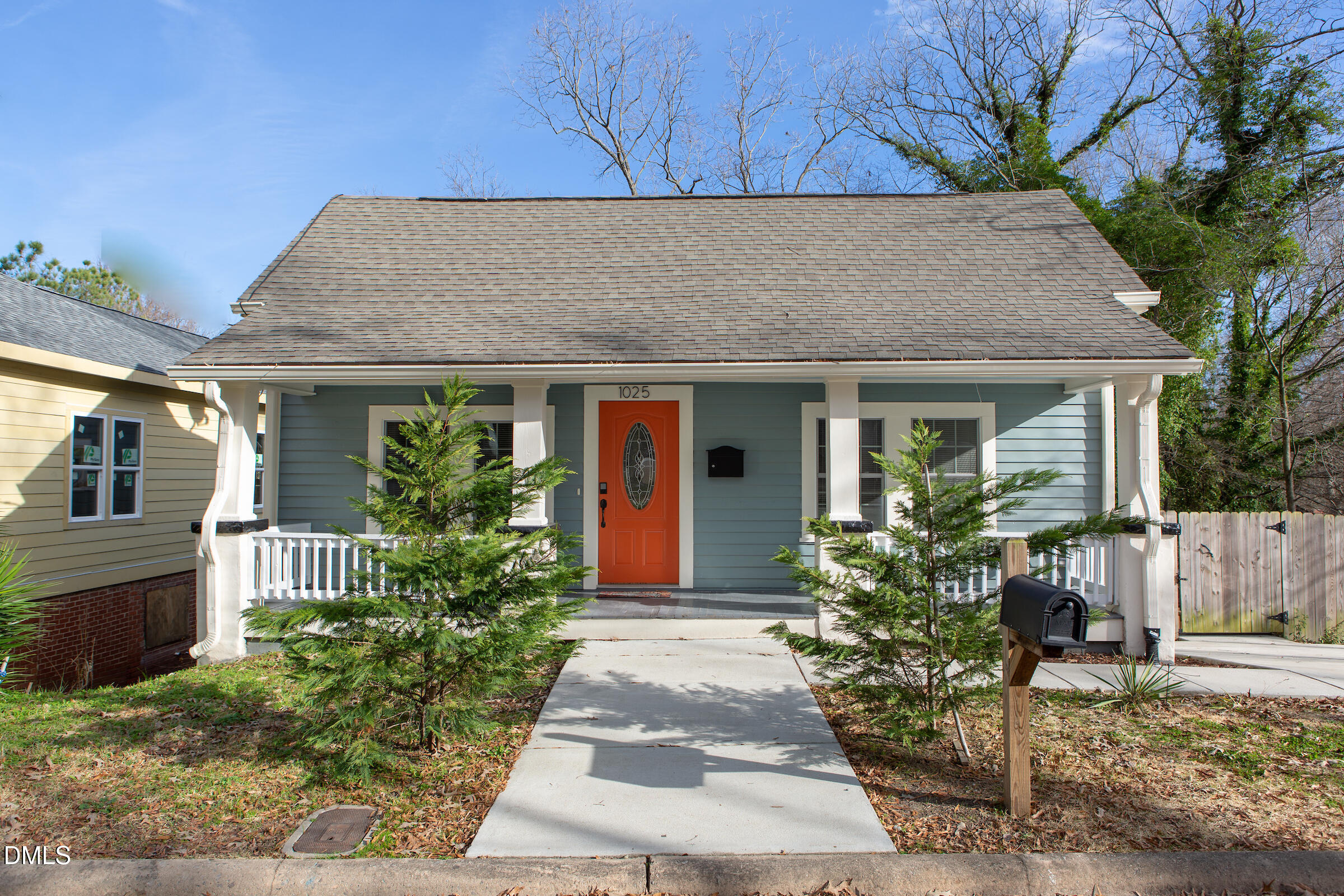 Blue-gray house with a covered white porch, orange-red front door, and concrete walkway between evergreen shrubs.