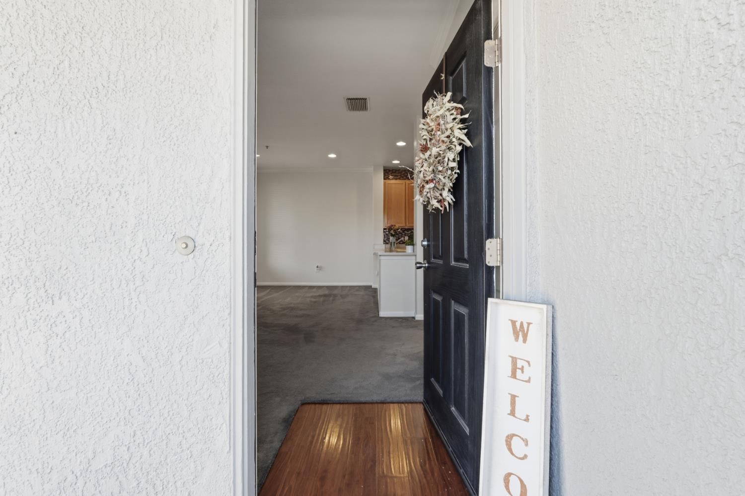 Open dark front door with a dried floral wreath and a welcome sign beside the entry.