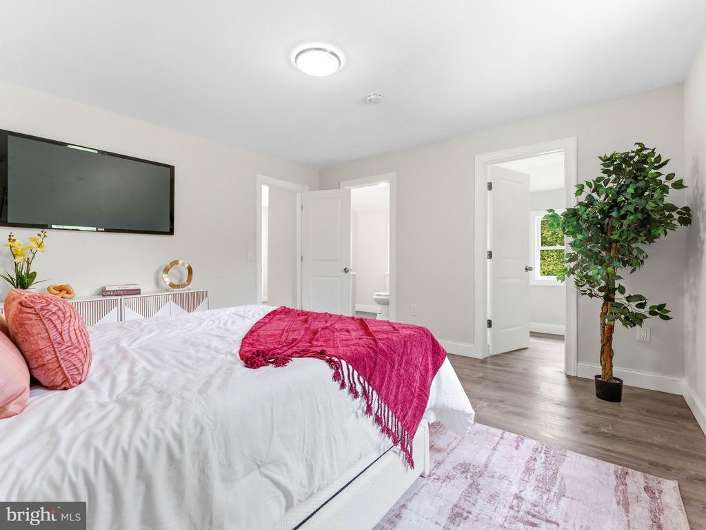 Staged bedroom with a white bed, vivid pink throw, wall-mounted television, and potted tree.