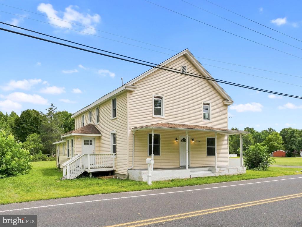Two-story beige house with a covered porch beside a road, photographed from the front corner.