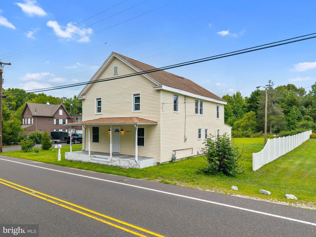 Two-story beige house with a covered porch beside a road and white side-yard fence.