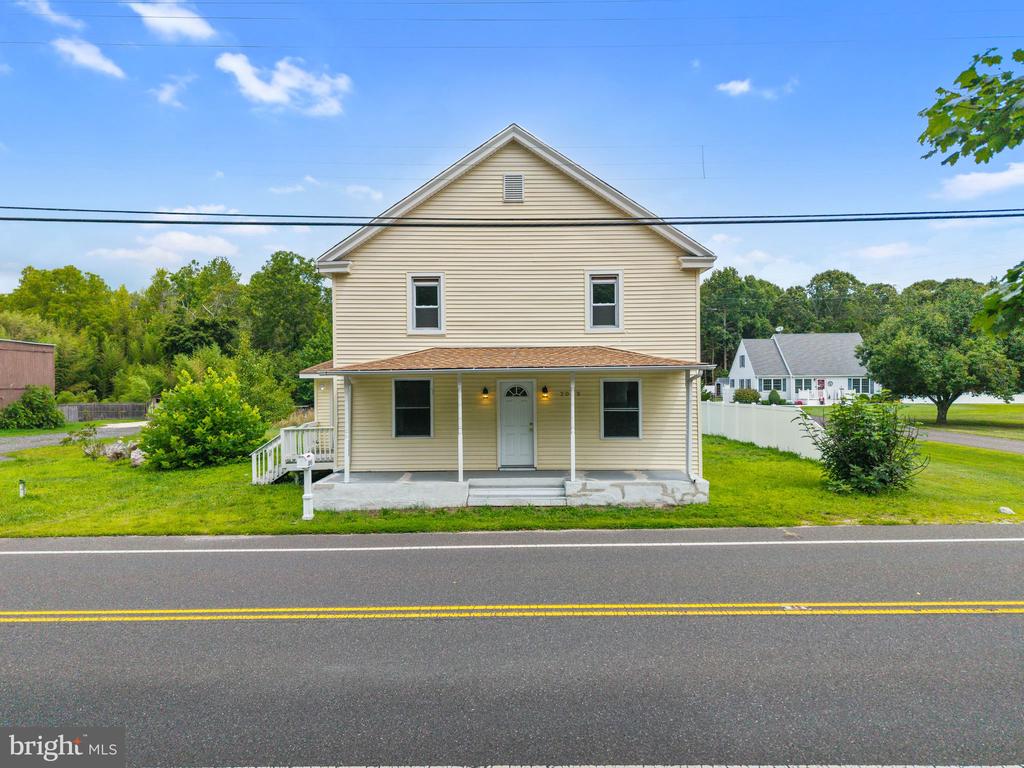 Two-story beige house with a covered porch, viewed from across a two-lane road.