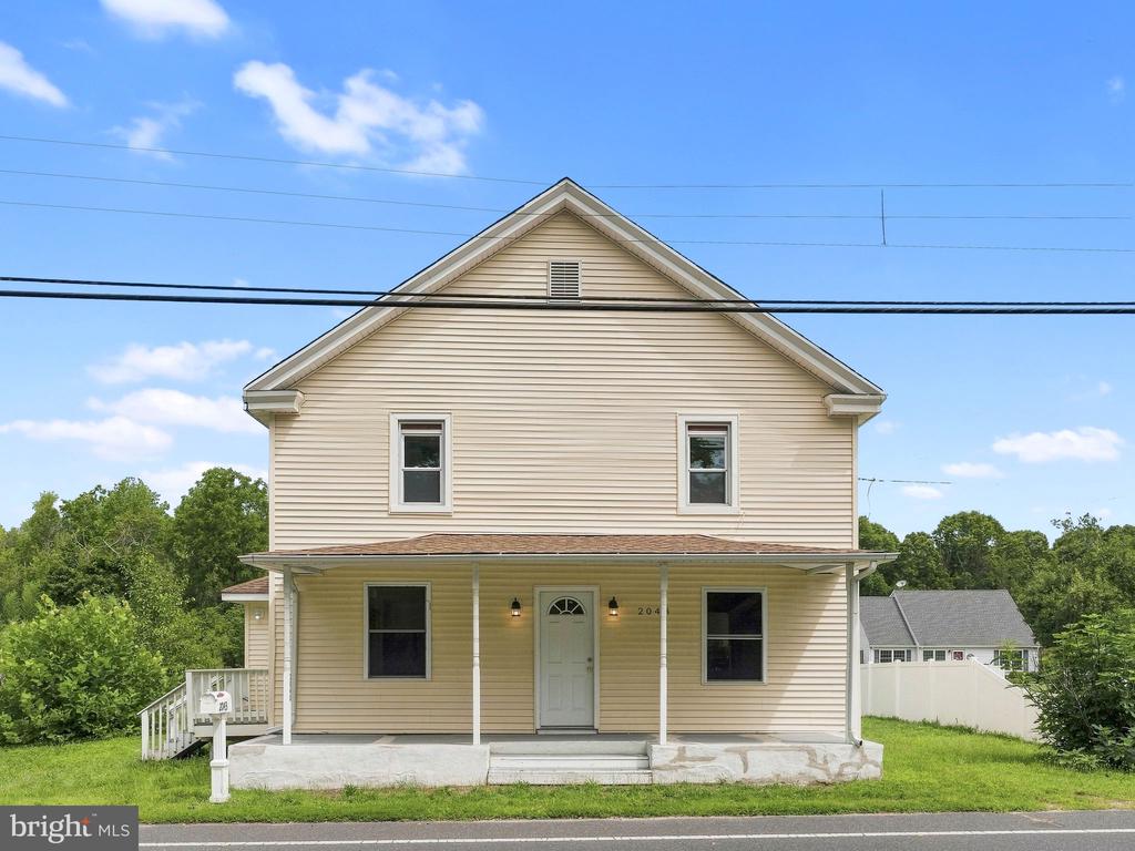 Two-story beige house with a covered front porch and lawn, viewed from across the road.