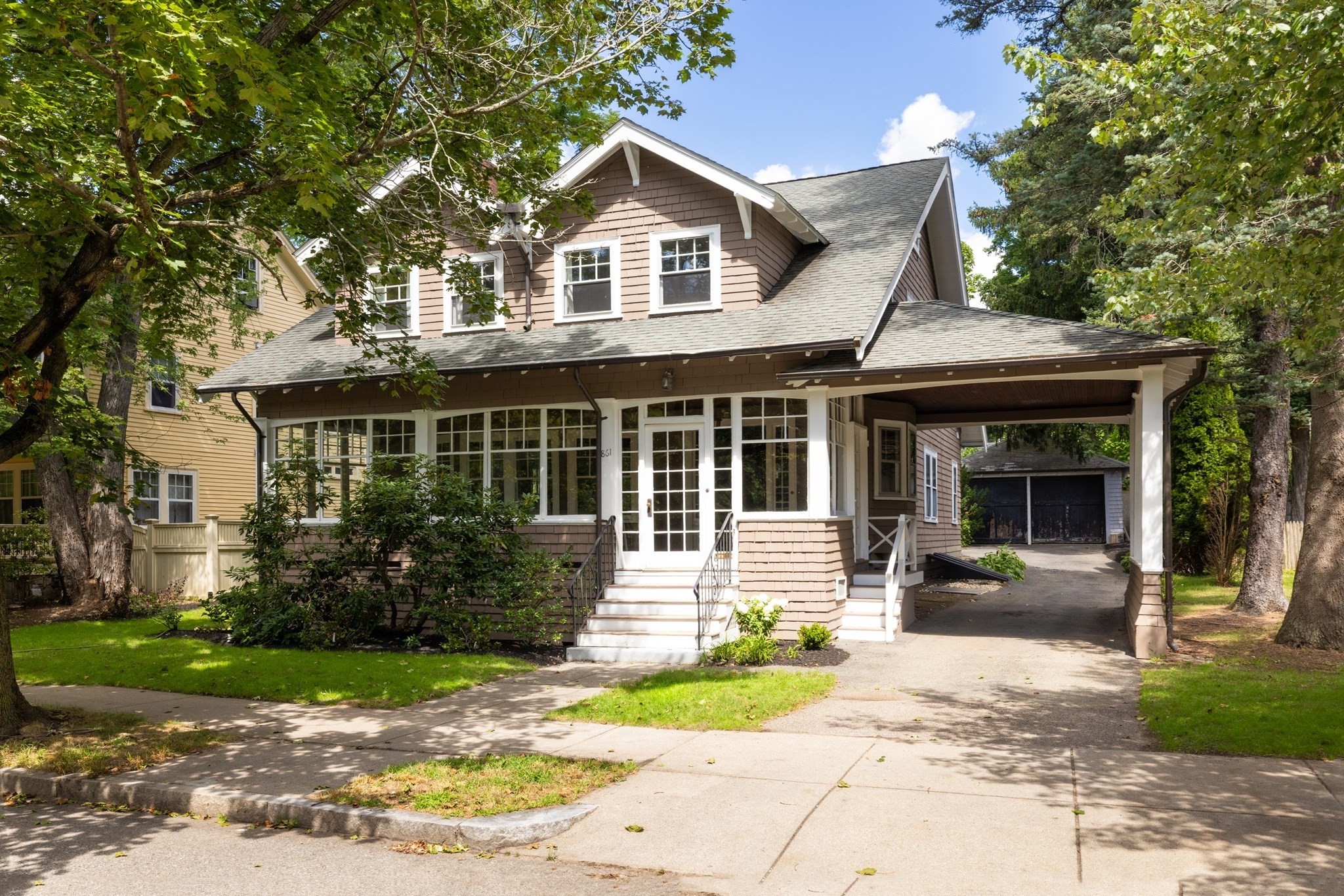 Two-story brown shingle-sided home with an enclosed porch and covered driveway, framed by mature trees.