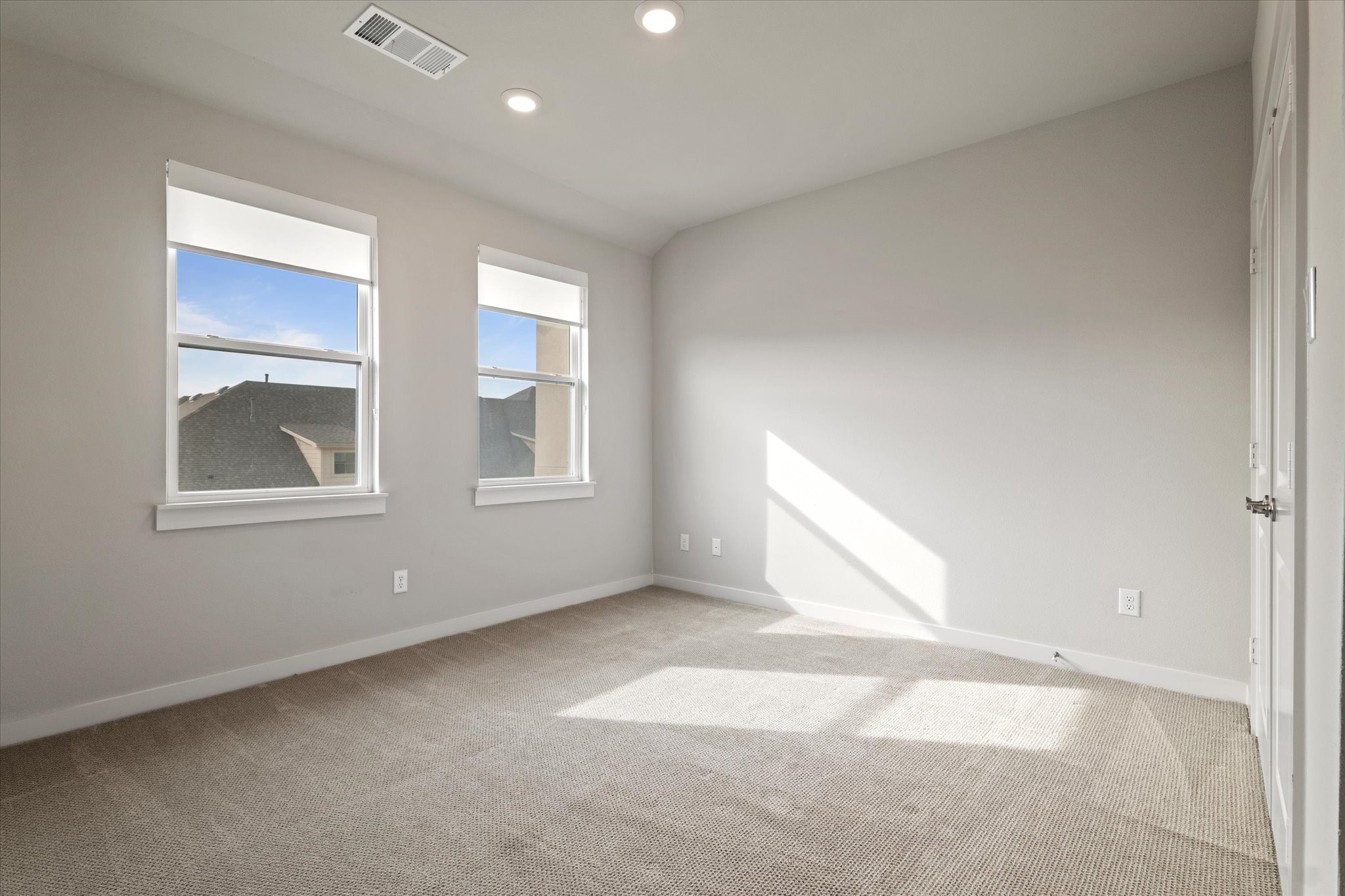 Empty bedroom with beige carpet, two windows, and sunlight across the floor.