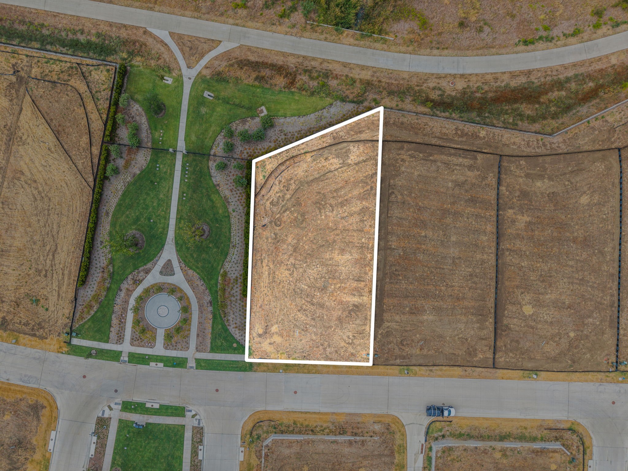 Aerial view of a vacant lot outlined in white beside a landscaped park and paved streets.