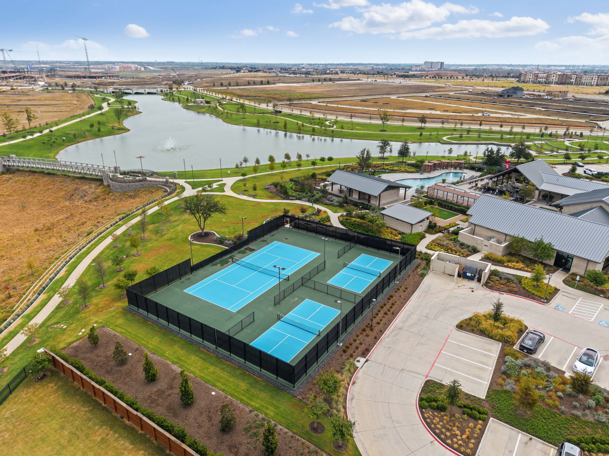 Aerial view of community tennis courts beside a pool, clubhouse, walking paths, and a lake.