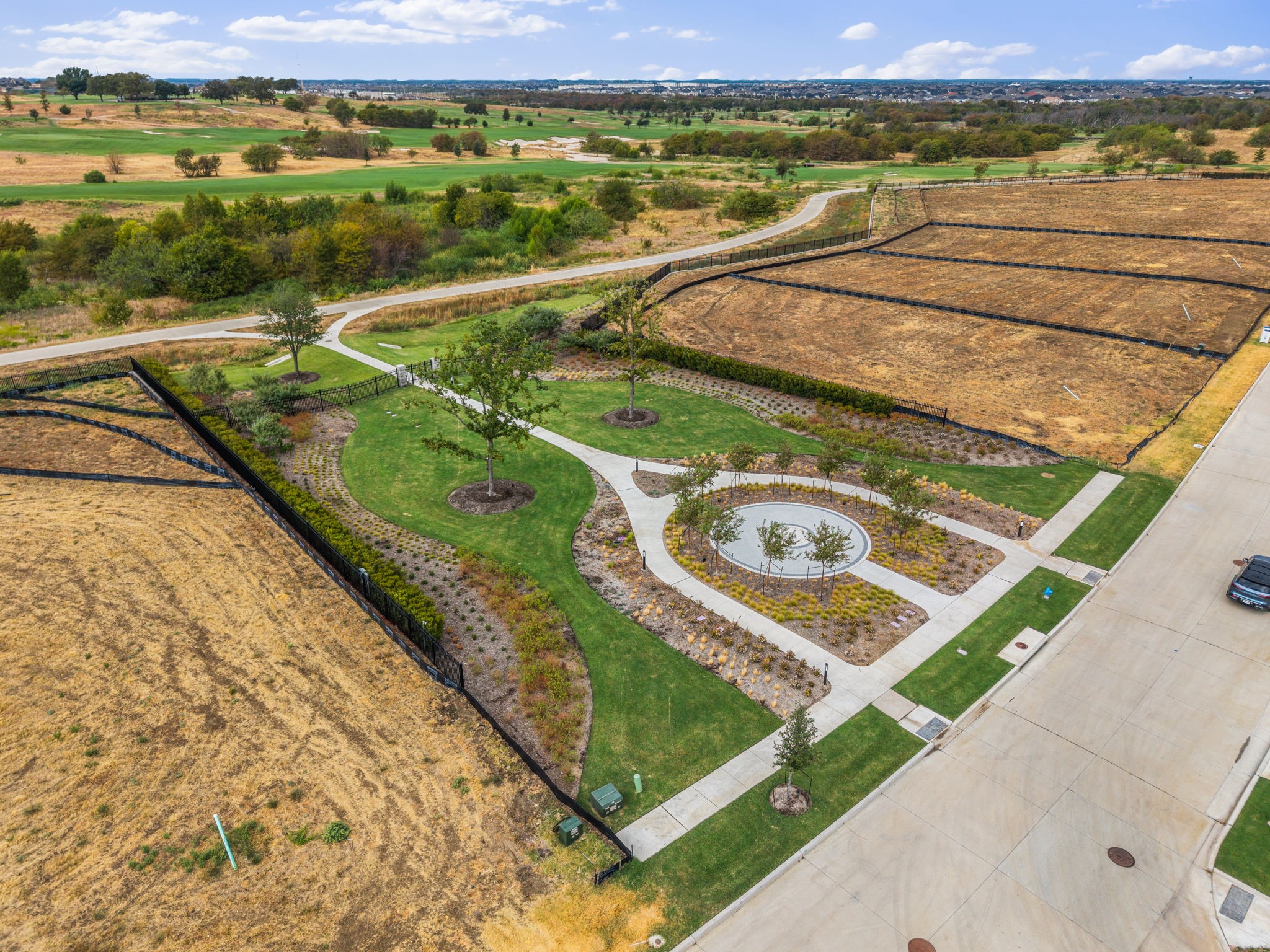 Aerial neighborhood greenspace with winding paths and a circular paved area beside dry vacant lots.
