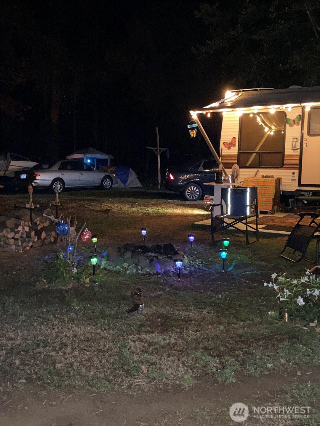 Camper trailer and cars beside a tent in a wooded yard lit by string and garden lights.