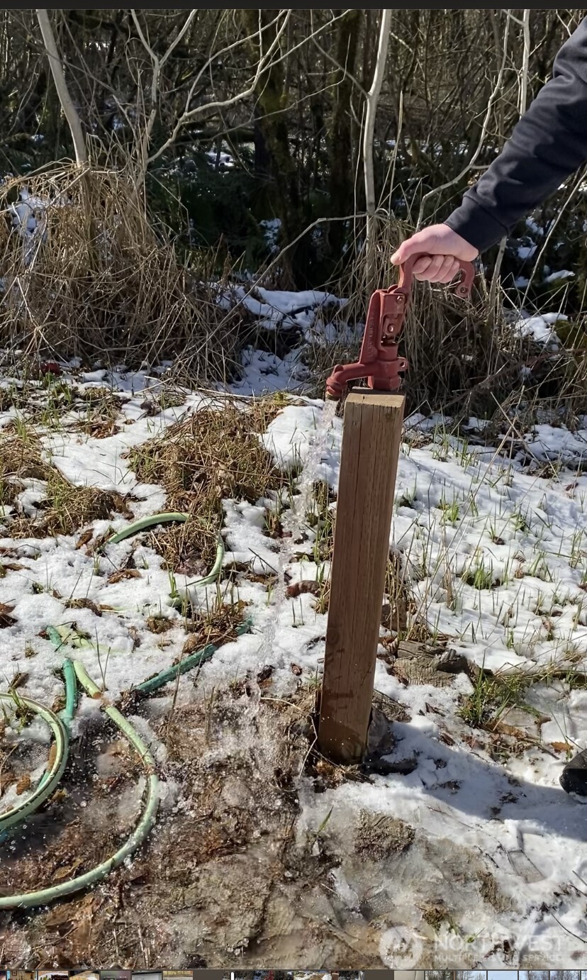 Outdoor hand pump running water onto snowy ground beside a green hose.