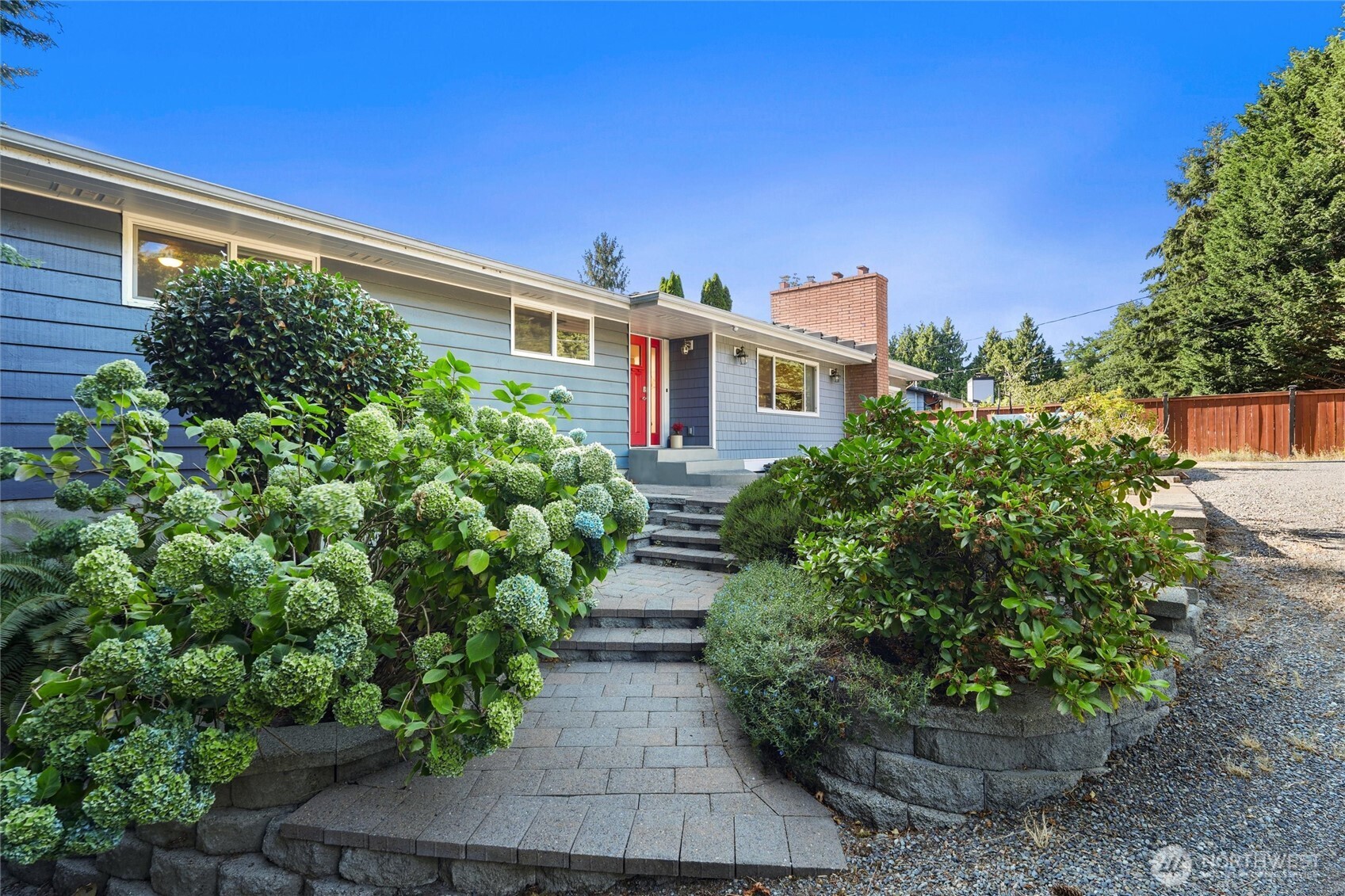 Blue single-story house with a red front door, brick chimney, and flower-lined stone walkway.