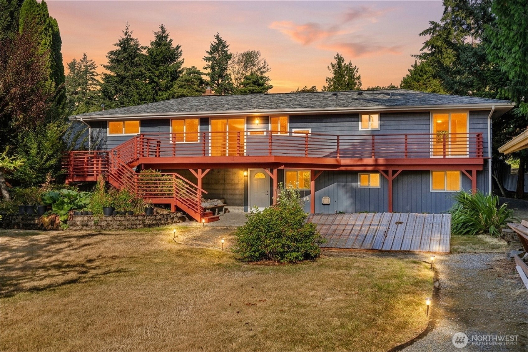 Gray split-level house with a long red-railed deck and exterior stairs, warmly lit at dusk.