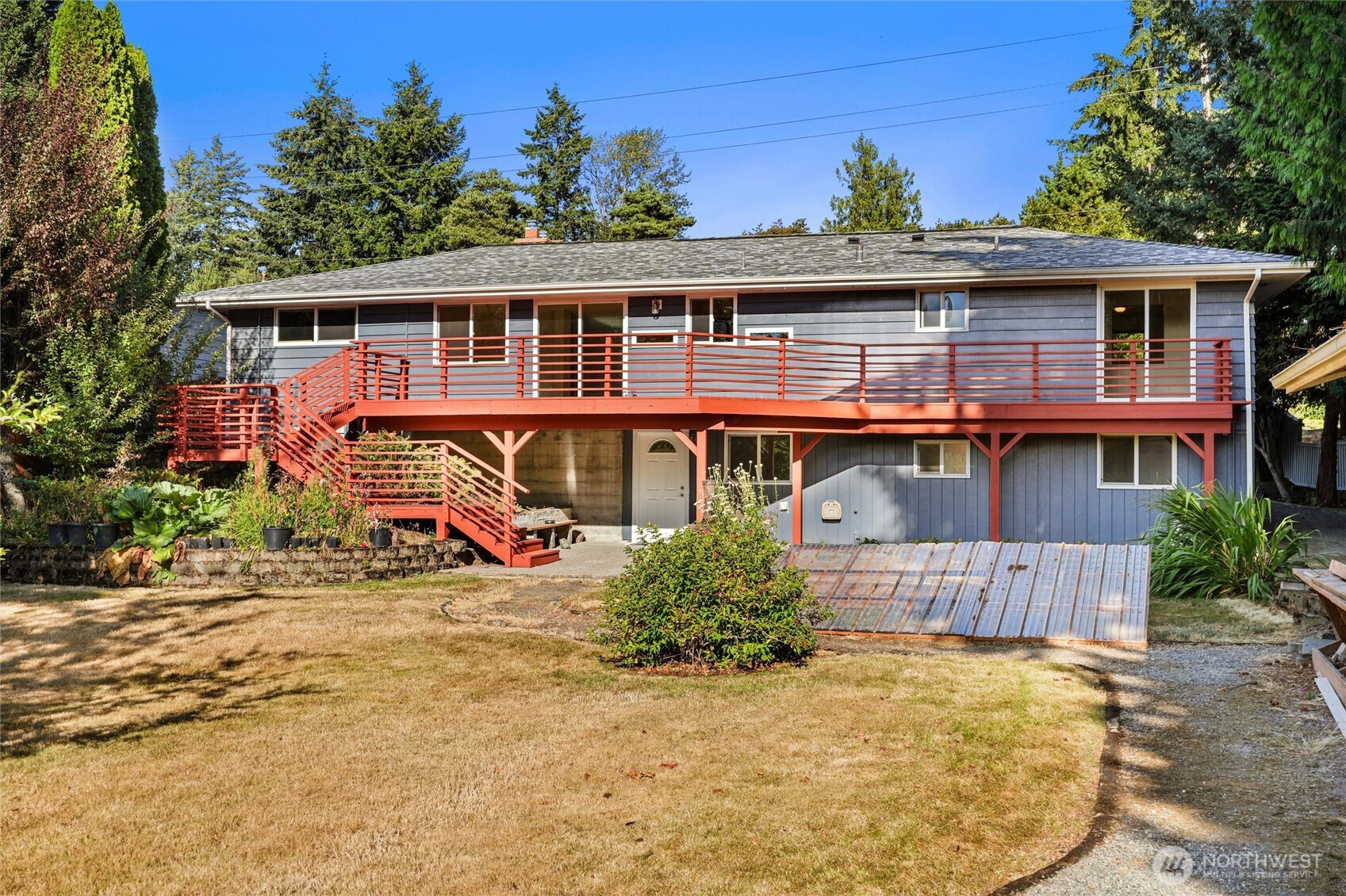 Rear of a gray split-level house with a long red-railed deck and stairs above a dry lawn.