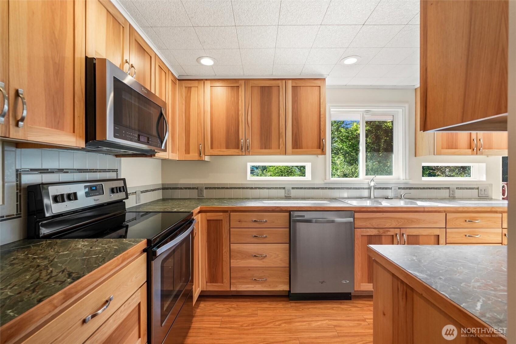 Kitchen with honey-toned wood cabinets, dark stone counters, stainless-steel dishwasher, and windows overlooking greenery.