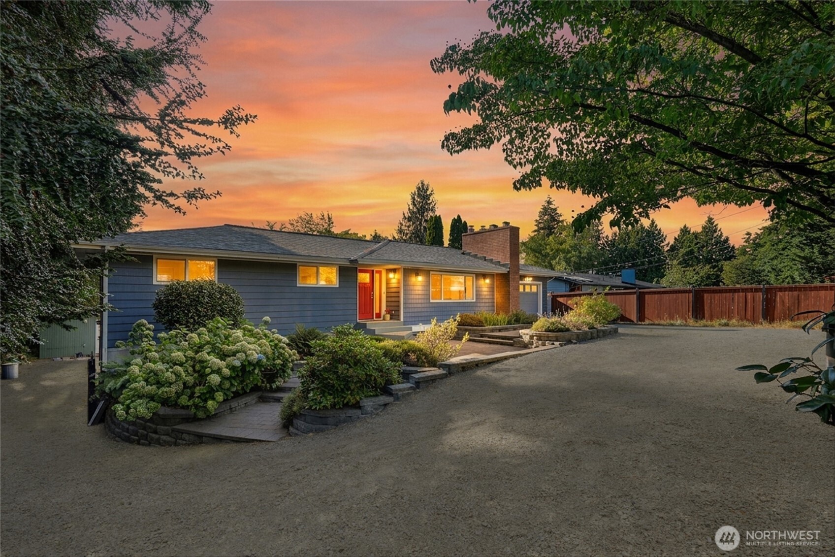 Single-story blue-gray house with a red door, landscaped entry, and glowing windows beneath an orange sunset.
