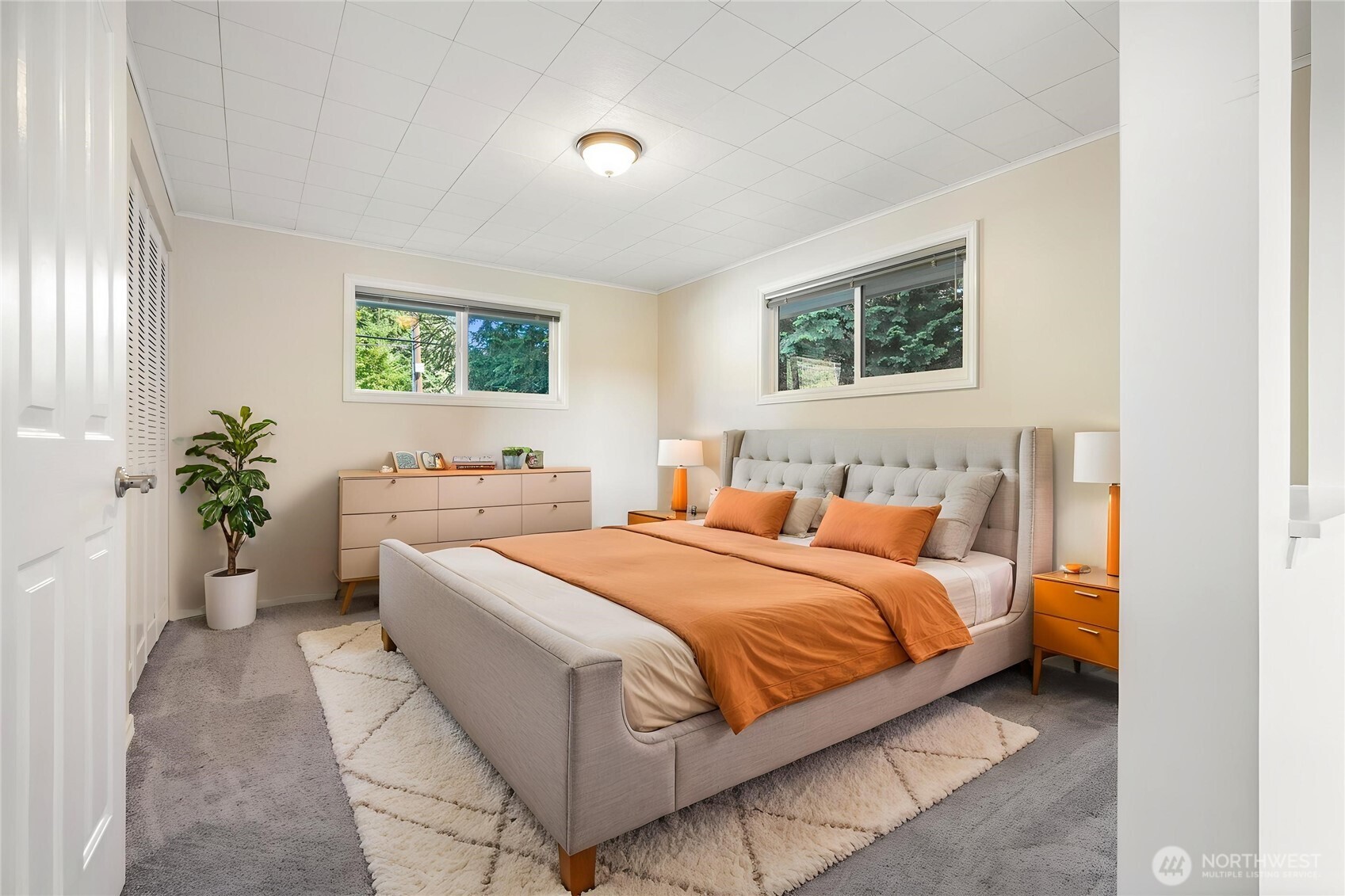 Staged bedroom with a large gray upholstered bed, rust-orange bedding, and two windows.