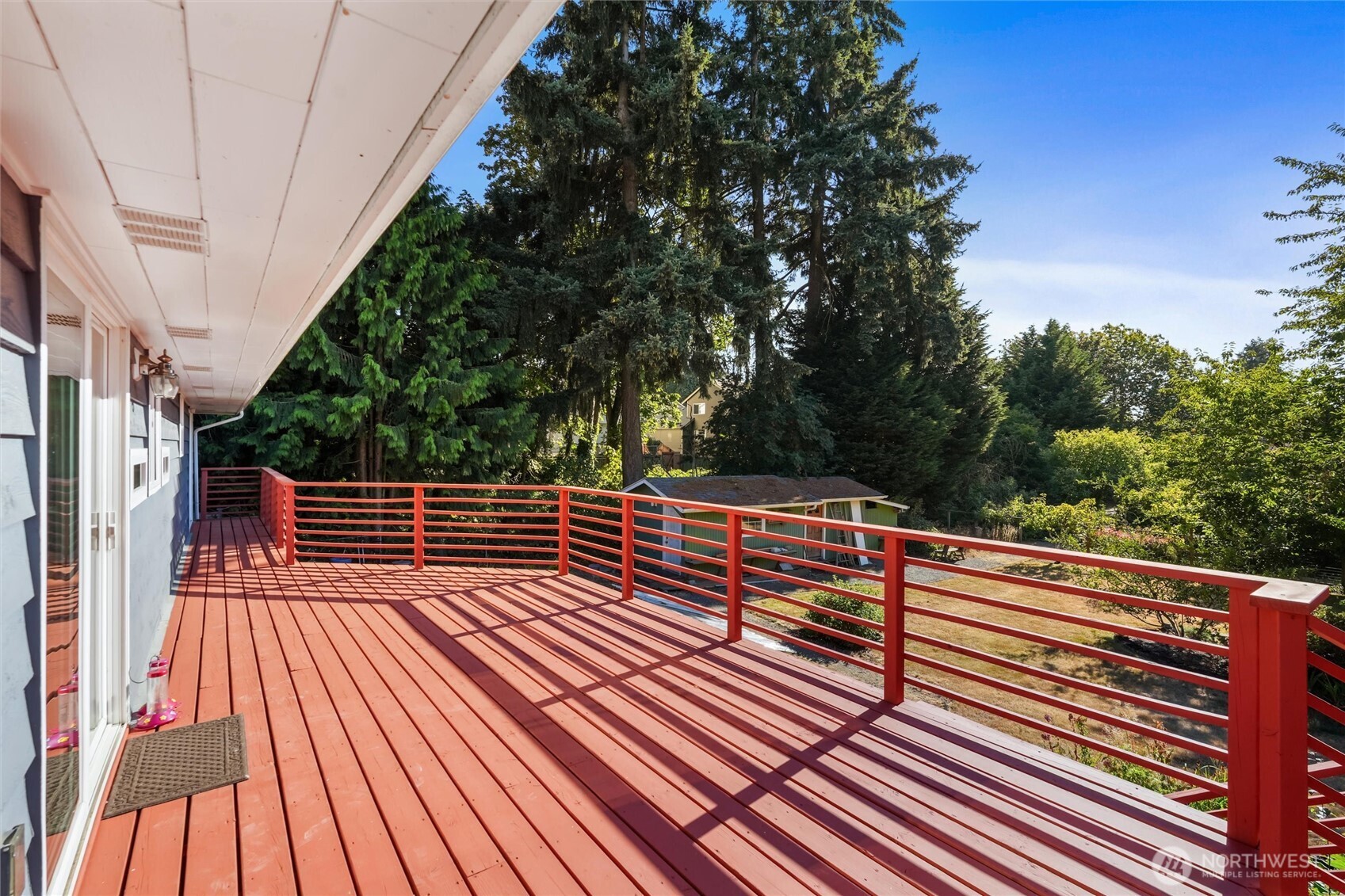 Red wooden deck with matching railings overlooking tall evergreen trees and neighboring yards.
