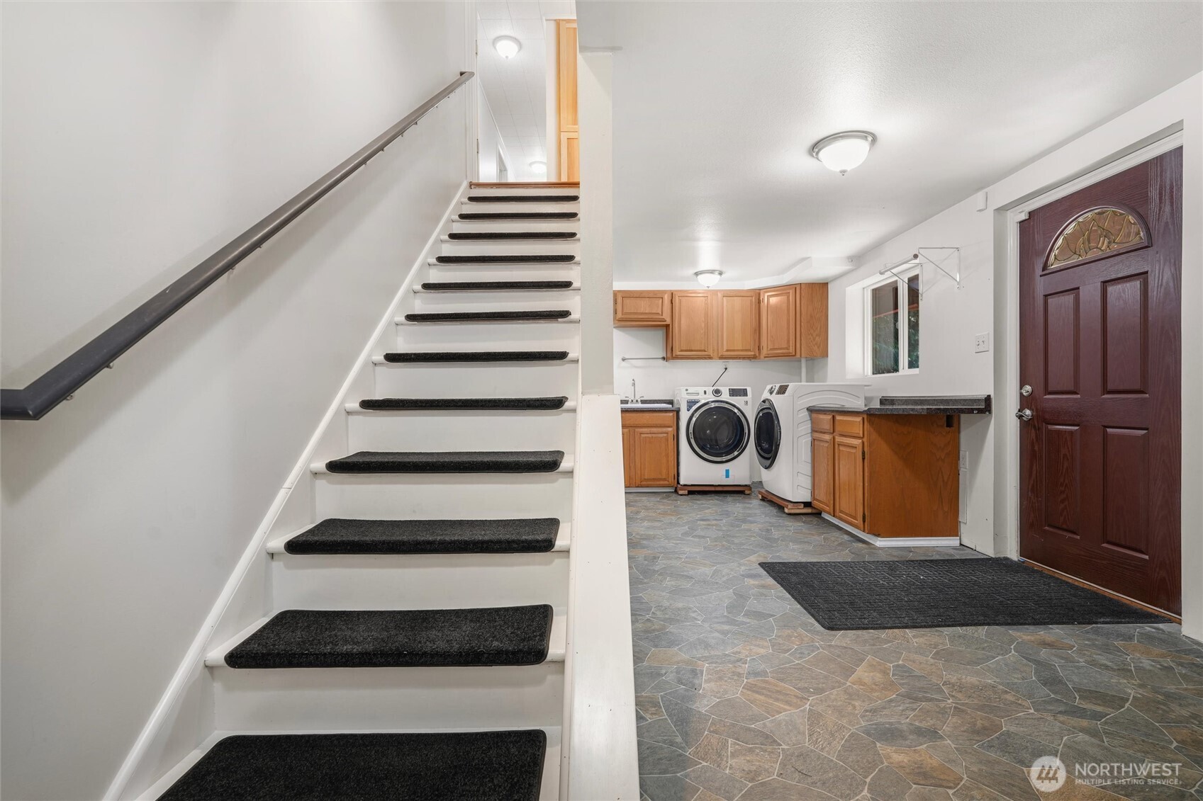 Entry and laundry area with carpeted stairs, wood cabinets, washer and dryer, and a dark red front door.