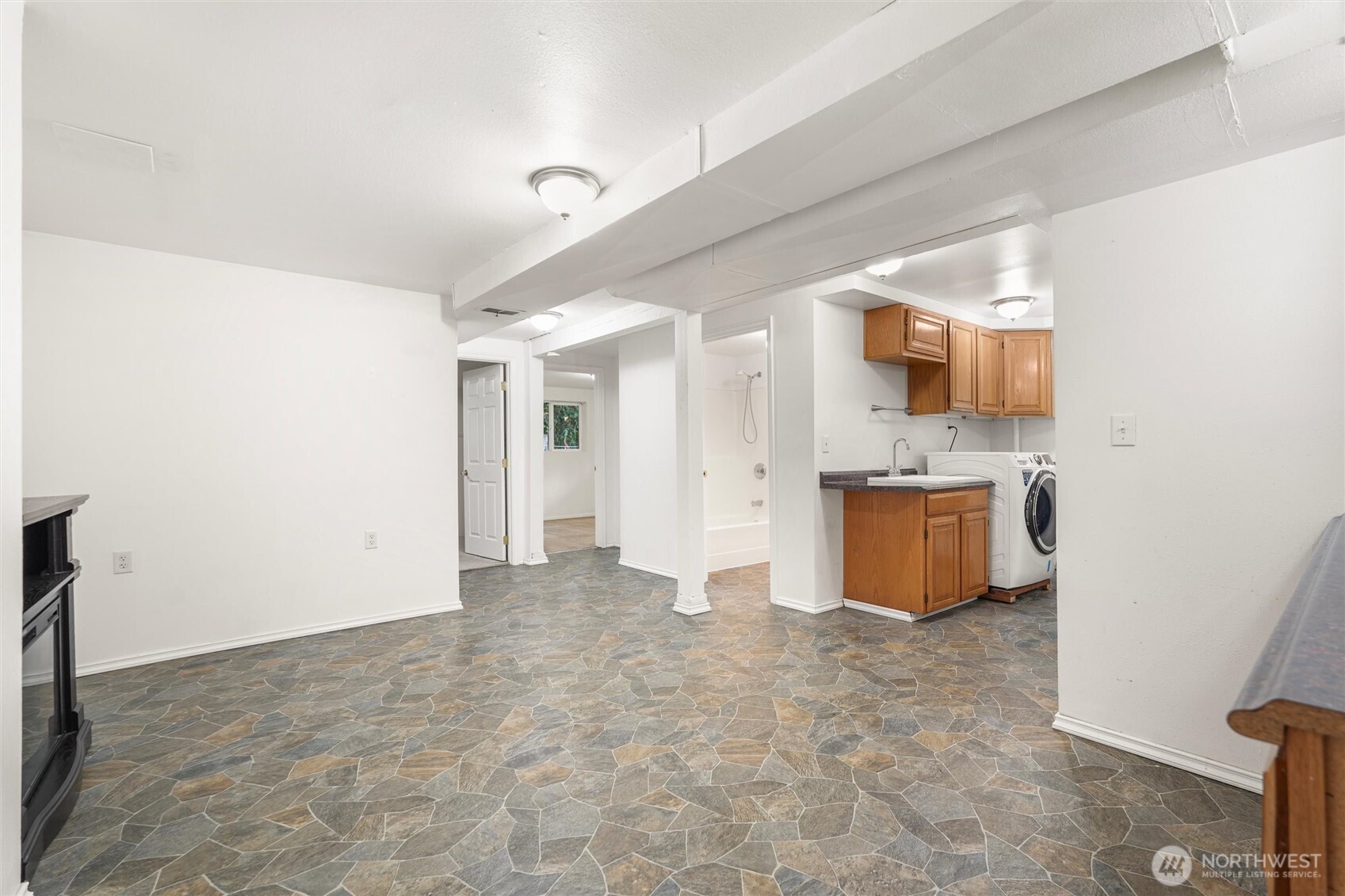 Basement laundry area with wooden cabinets, washer, bathtub, and multicolored stone-pattern flooring.