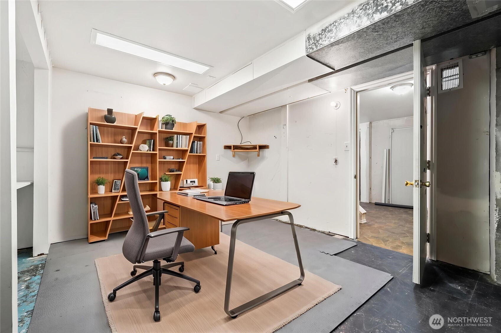 Home office with a wood desk and shelving, gray swivel chair, and doorway to an adjoining room.