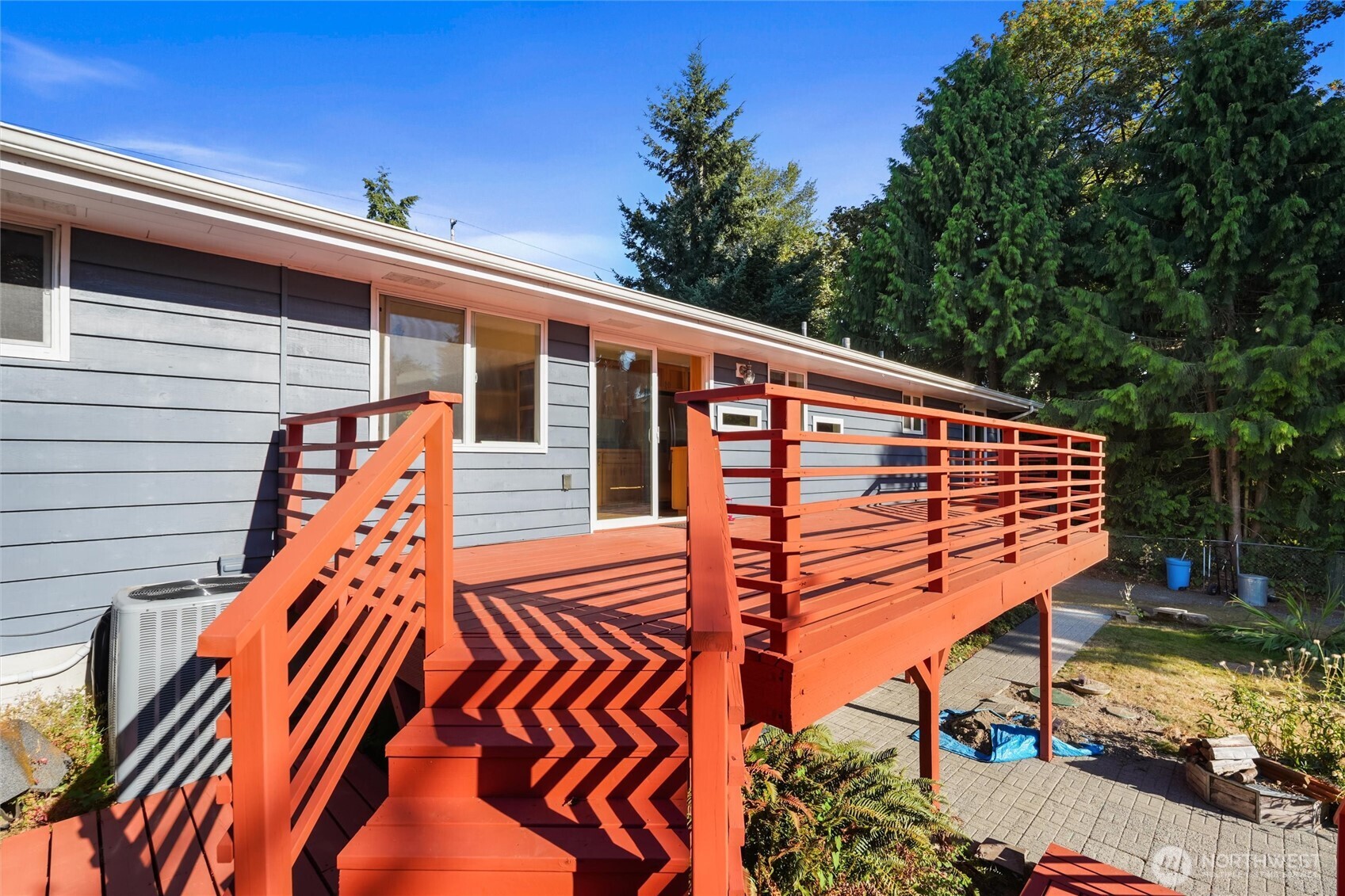 Gray-sided house with a large elevated red-orange deck, broad stairs, and mature evergreens.