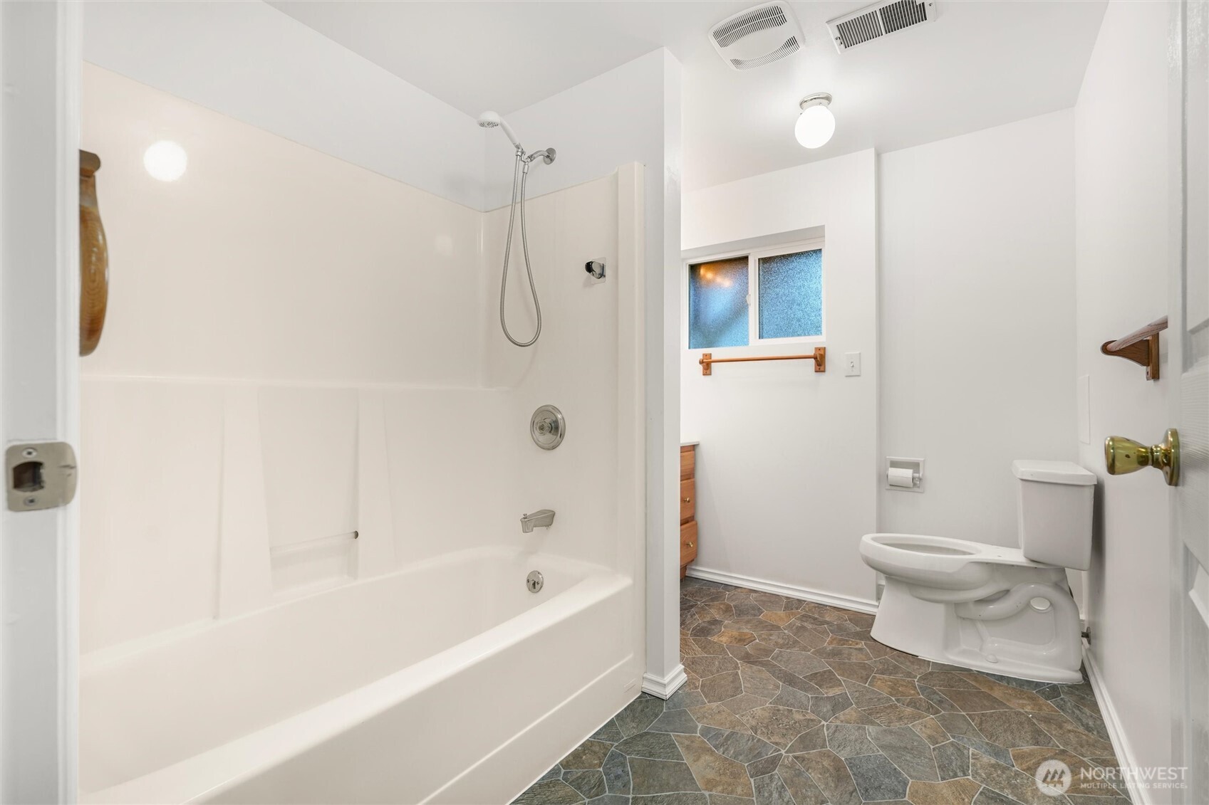 Bathroom with a white tub and shower, toilet, frosted window, and stone-pattern tile floor.