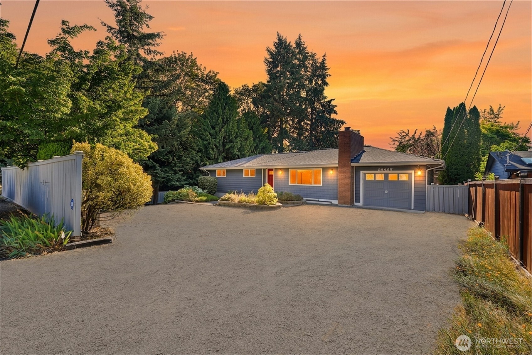 Single-story gray home with brick chimney and garage beyond a broad gravel driveway at sunset.