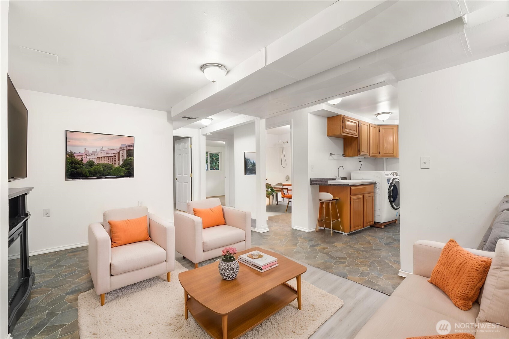Living room with pale seating, orange pillows, stone-pattern flooring, and an open kitchen.