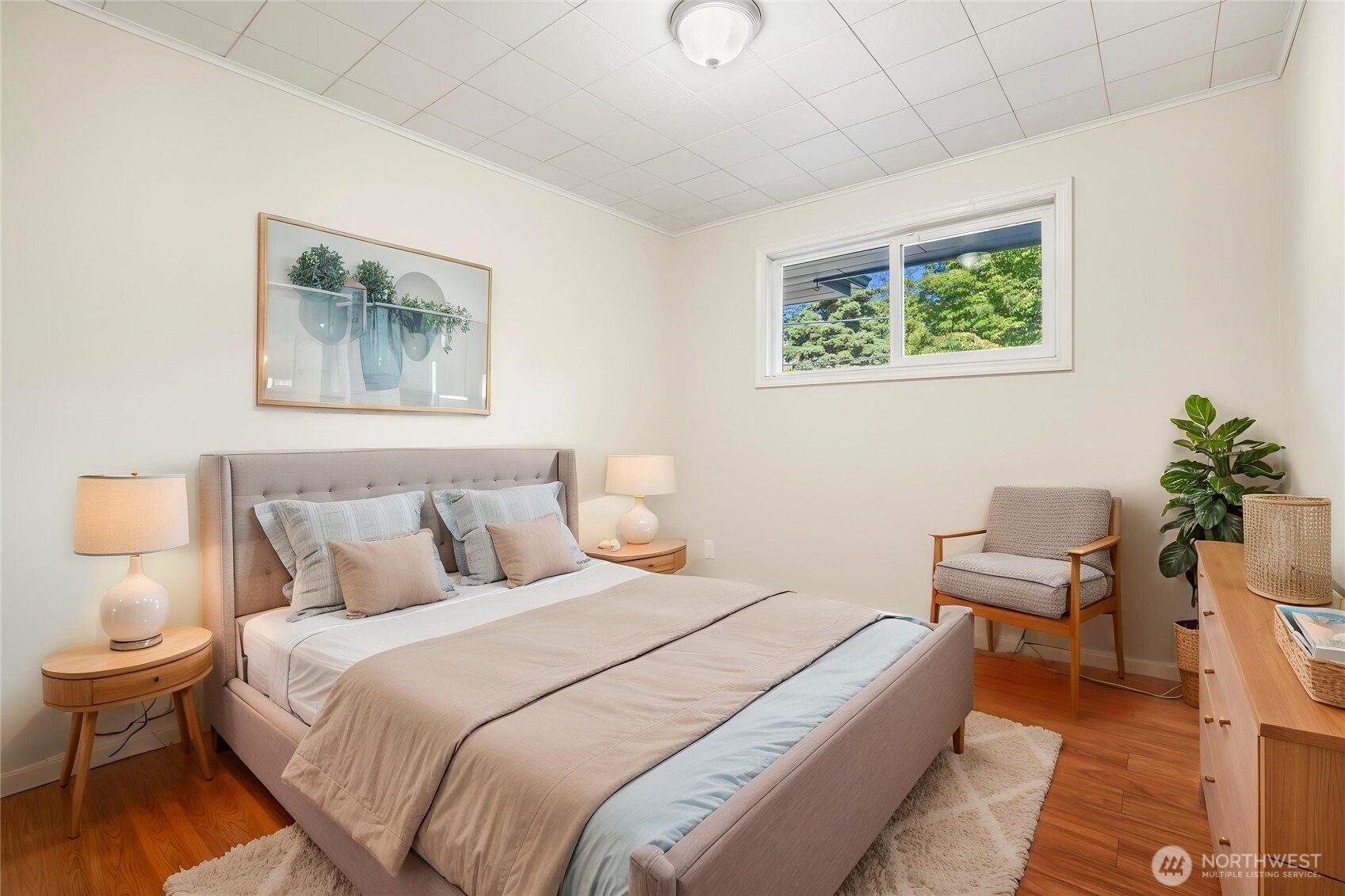Staged bedroom with a neutral upholstered bed, wood furnishings, and a window overlooking trees.